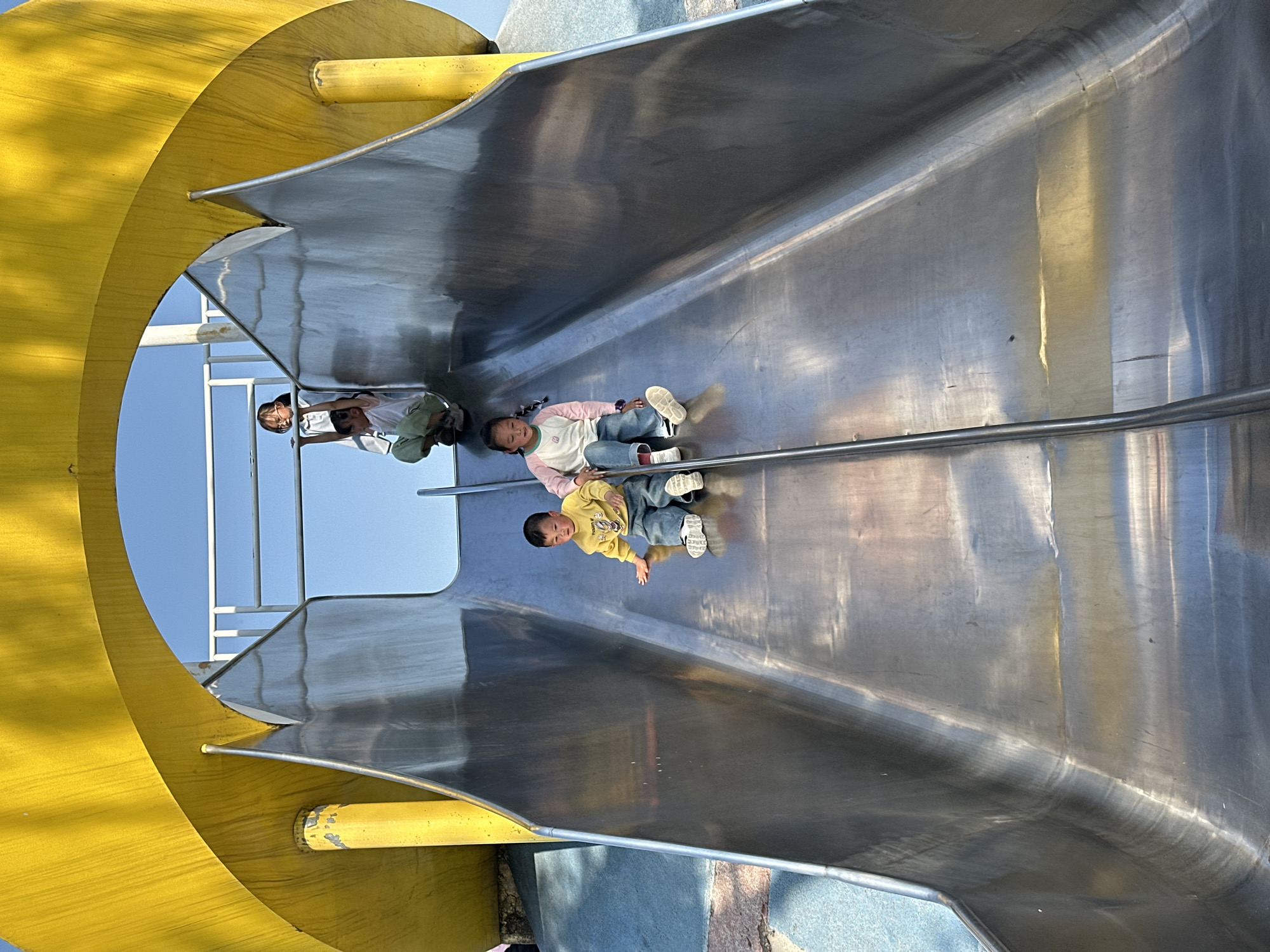 Three children are on a playground slide, presumably in Jiangjin, China. One child is wearing a yellow sweatshirt, another is wearing a light pink top with jeans, and a third child is at the top of the slide. The slide is metal with a yellow support structure. The sky is visible above the children at the top of the slide.