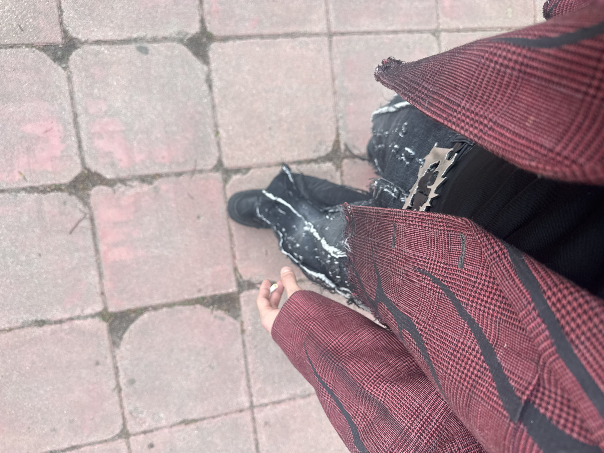 An overhead shot shows a person standing on a brick sidewalk. The person wears black boots, heavily distressed black jeans with white splatters, a black garment, and a red and black plaid blazer. A ring is visible on the person's left hand. The background is a pedestrian zone paved with reddish-grey paving stones. This area shows some wear and tear, with visible cracks and weeds growing in the gaps between the stones. The location is likely a public space in Sofia, Bulgaria, based on the type of paving stones commonly used in Bulgarian cities.