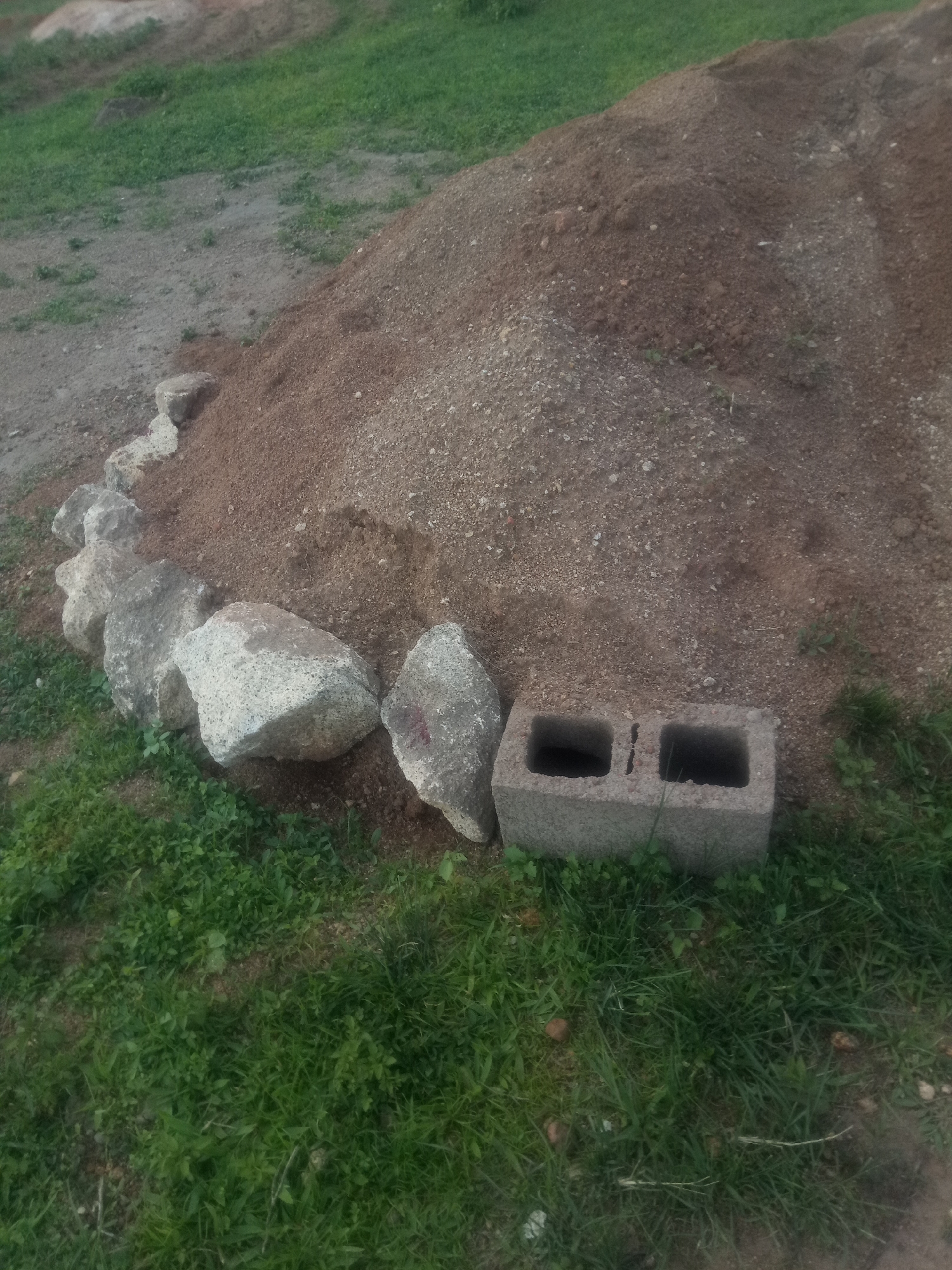 The image shows a pile of dirt with some rocks and a cinder block in the foreground. The dirt pile is large and appears to be freshly dug or dumped. Several large, gray rocks are arranged in a semi-circle along the edge of the dirt pile, bordering a patch of green grass. A single gray cinder block, with two hollow rectangular openings, sits on the grass near the rocks. The background consists of more dirt and grass, suggesting an outdoor, possibly construction or landscaping, setting. The lighting and shadows indicate it is daytime, but the weather is not discernible. There is no visible text in the image.