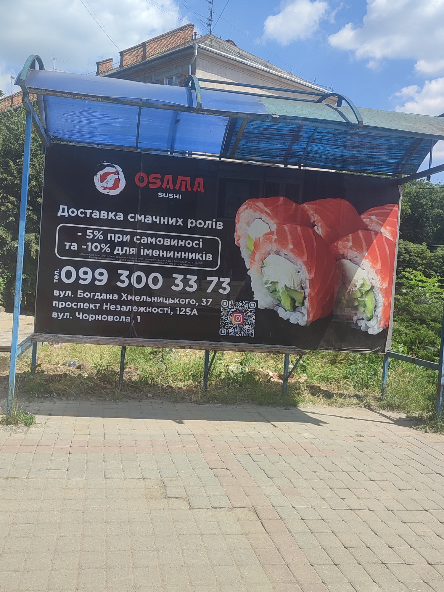The image shows an outdoor banner advertising "OSAMA SUSHI" in Chernivtsi, Ukraine. The banner features images of sushi rolls and text in Ukrainian. Key information visible on the banner includes a phone number "099 300 33 73", and addresses: "вул. Богдана Хмельницького, 37" (Bohdana Khmelnytskoho Street, 37) and "проспект Незалежності, 125А вул. Чорновола, 1" (Independence Avenue, 125A Chornovola Street, 1). The banner is attached to a metal frame structure with a blue translucent roof, likely a bus stop or shelter. The background shows a building with a brick facade and a blue sky with clouds. The ground is paved with light-colored rectangular tiles, with some grass and dirt visible. There are no people visible in the image.