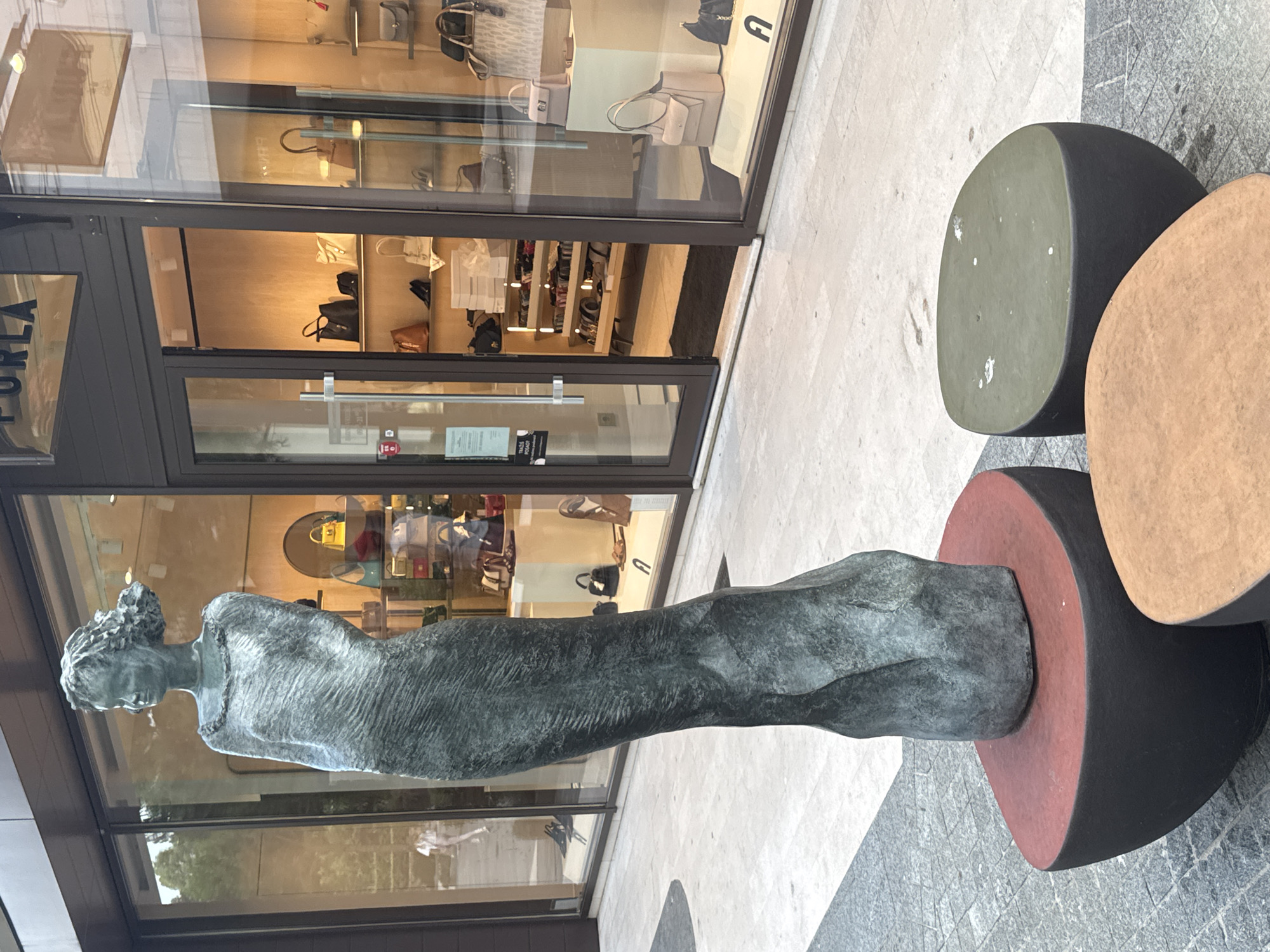 This is a vertical, eye-level, outdoor shot of a bronze statue of a woman in front of a storefront. The statue depicts a woman from the chest up, with her head tilted slightly down and her hair styled in an updo. Her arms are not visible. The statue has a textured, weathered appearance.
Behind the statue is a storefront with large glass windows, revealing the interior of a boutique. The store appears to be selling handbags and possibly shoes, with various items displayed on shelves and mannequins. The interior is brightly lit and has wooden accents. The word "FURLA" is visible on a sign on the left side of the storefront.
To the right of the statue, there are three large, abstract, circular sculptures. The topmost is a pale green, the middle one is a light brown, and the bottom one is dark red. These sculptures are placed on a textured, light-colored paved surface. The ground in front of the storefront is a mix of grey cobblestones and lighter paving stones.
The lighting suggests it is daytime, possibly late afternoon or early evening, as there are some shadows, but the scene is well-lit. The weather appears to be clear. The presence of a retail store and outdoor sculptures suggests an urban or commercial setting. There are no people clearly visible in the statue or the storefront, though reflections in the window might obscure some details.