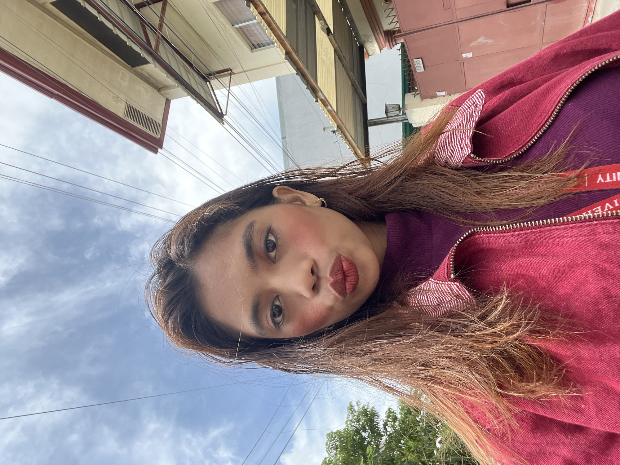 A medium shot of a woman is pictured from a low angle. The subject has fair skin, brown hair, and wears a purple top with a red jacket. The background features a cloudy sky, green trees, and city buildings. Power lines are visible in the sky. The setting suggests a location in Davao City, Philippines.