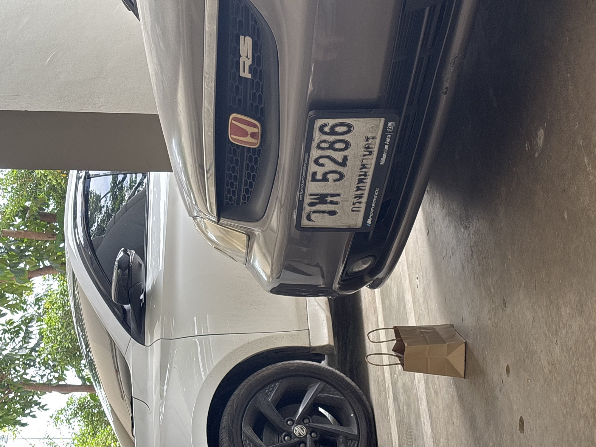 The image shows two cars parked in a garage in an unidentified location in Choeng Thale, Thailand. The car in the foreground is gray and displays a license plate that reads "วท 5286 กรุงเทพมหานคร." The emblem on the grill is an RS model. The car behind is white. A small brown paper bag sits on the floor between the two cars.