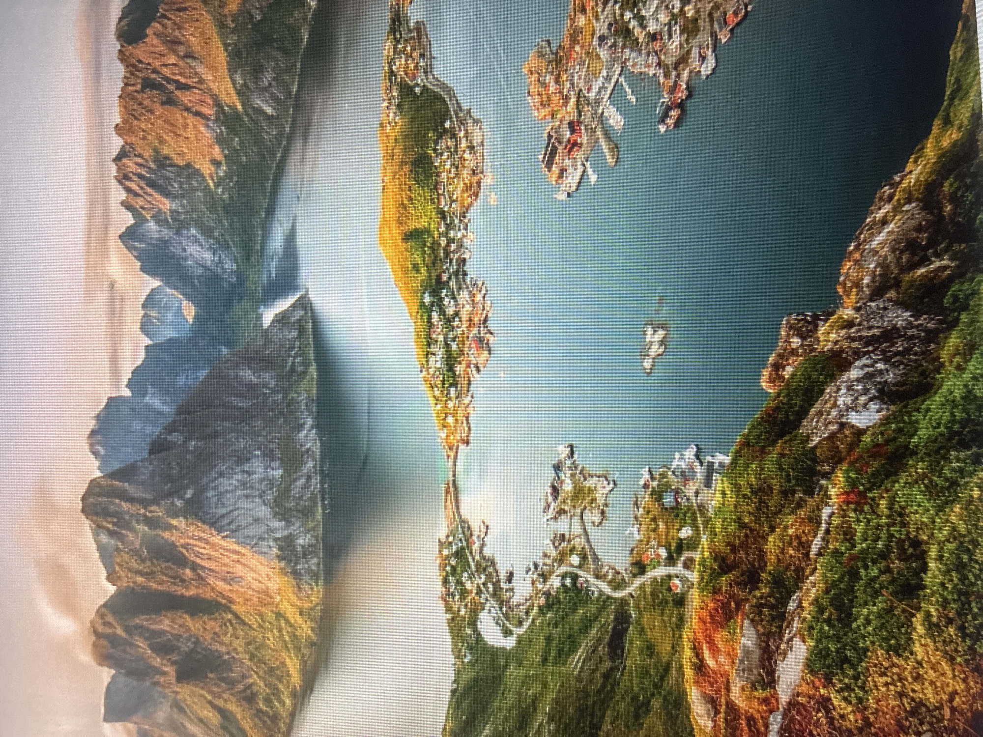 The image, captured from a high aerial perspective, showcases a dramatic mountainous coastal landscape resembling a fjord system. The scene is oriented vertically, with towering, rugged mountains dominating the left side. These mountains feature steep rock faces in shades of dark grey, brown, and ochre, interspersed with patches of green and reddish vegetation. Sunlight, appearing to be from a low angle (either early morning or late afternoon), casts a golden glow on some mountain slopes and creates long shadows.
On the right side, a vast expanse of dark blue-green water defines the fjord, dotted with numerous islands and narrow peninsulas. These landmasses are extensively developed with dense clusters of buildings, forming towns or settlements built right up to the water's edge. The structures, visible in various colors including white, red, and brown, are connected by winding roads and paths. Several docks or harbors with smaller boats are discernible along the shoreline.
No people or specific activities are taking place; the image presents a static view of a populated natural environment. The weather appears clear and calm, with no clouds visible and the water surface relatively smooth. There is no visible text in the image. The geographical features, particularly the deep, narrow inlets and steep, rugged mountains, are characteristic of fjord landscapes.