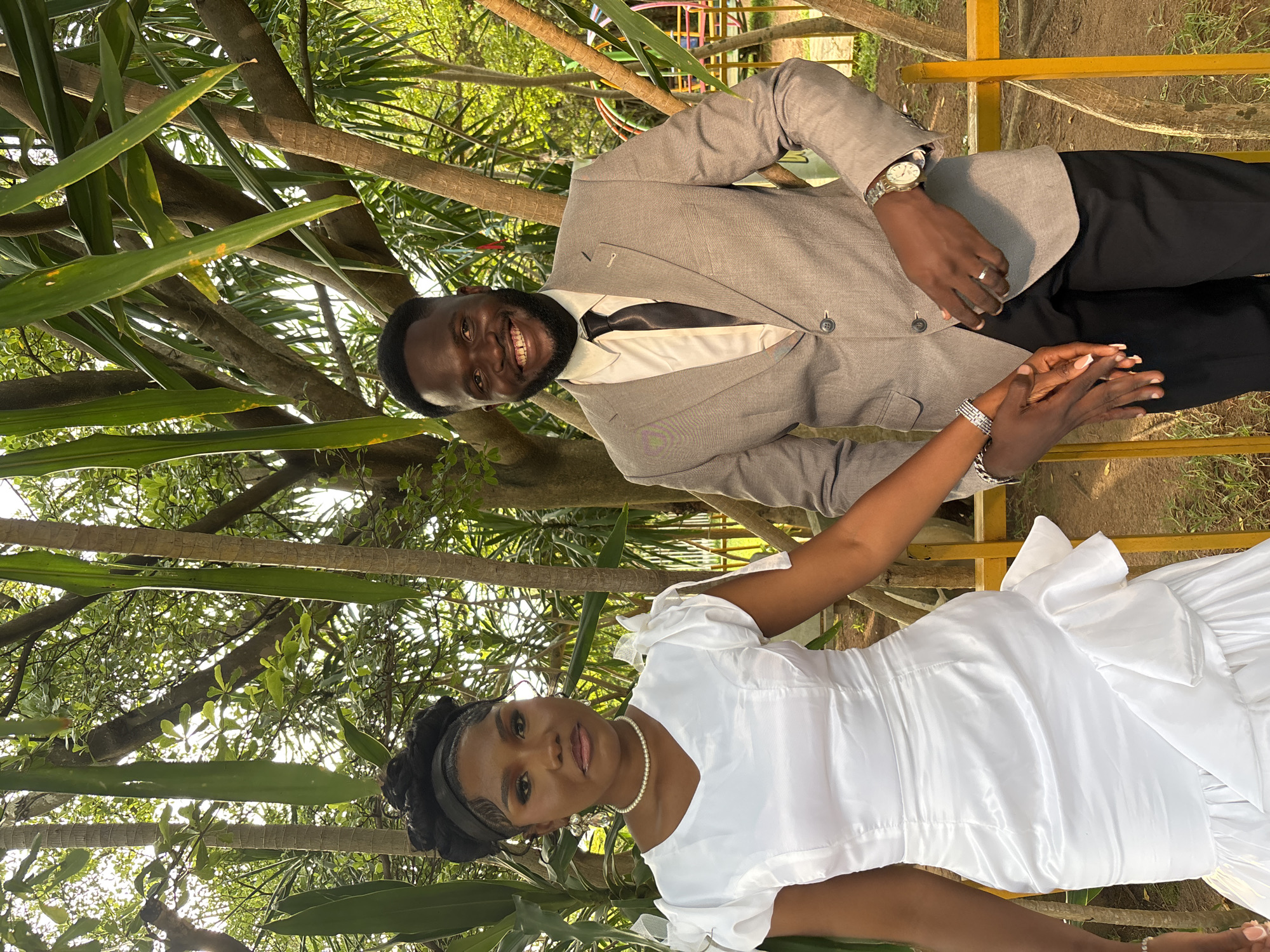 A wedding photograph is depicted in the city of Barikin Gidar Kanat, Nigeria. A Black couple, likely the bride and groom, stand outdoors, holding hands. The bride is on the left, wearing a white, knee-length dress with ruffled detailing at the hem and puffed sleeves. She has her hair styled in an updo and is adorned with a pearl necklace and dangling earrings. The groom stands to her right, dressed in a light grey suit jacket, a white shirt, and a black tie. He has a beard and is smiling at the camera. They are positioned in front of a backdrop of lush greenery, with trees and foliage surrounding them, suggesting a garden or park setting. A yellow railing is visible in the foreground.