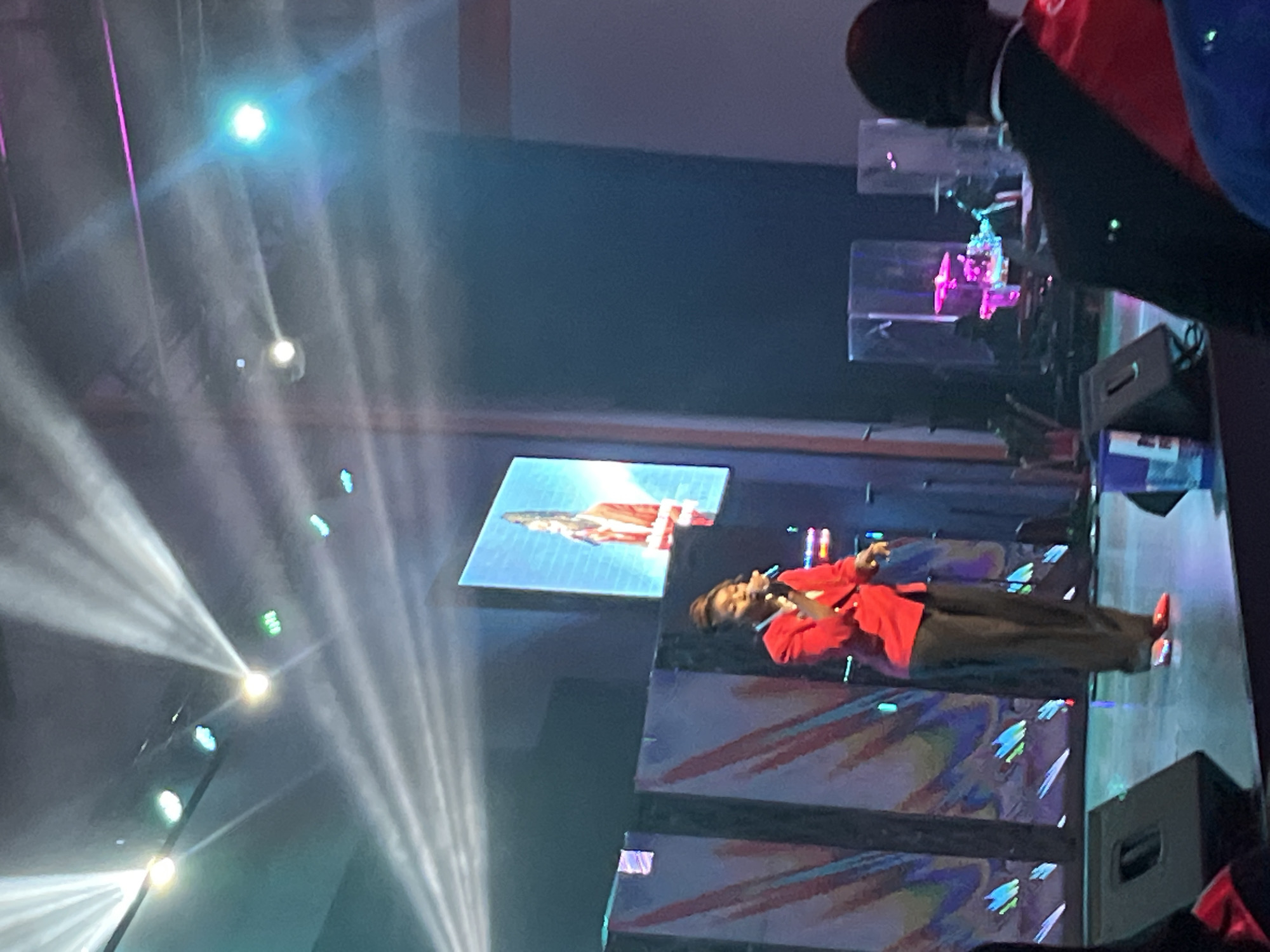 A woman in a red blazer and black pants stands on a stage, holding a microphone and singing. She has her hair pulled back in a bun. A large screen behind her displays a similar image of her singing. The stage is lit with bright spotlights, and there are speakers and other equipment visible. In the background, there are translucent structures with colorful lights. The scene appears to be a performance or event in Abuja, Nigeria.