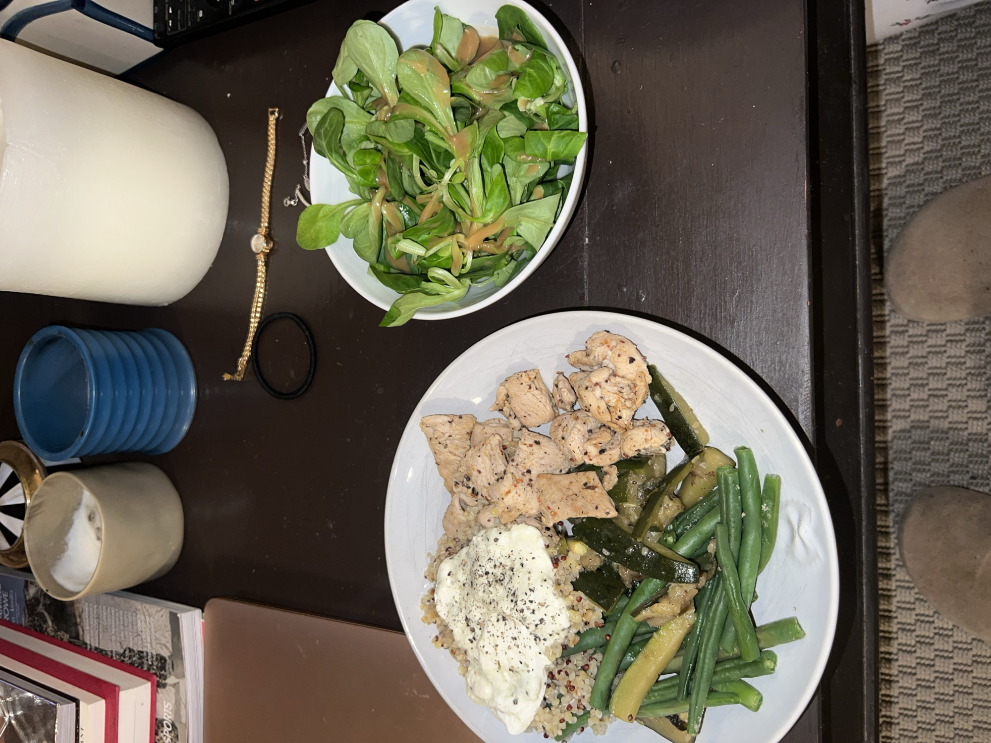 The image shows a table with two bowls of food. One bowl contains a green salad with dressing. The other bowl contains chicken, green beans, zucchini, quinoa, and a white sauce. There are also other objects on the table, including a white candle, a blue vase, a cup, books, a laptop, a gold watch, and a hair tie. A portion of a person’s feet wearing tan-colored slippers is visible on the right side of the image. The location is not identifiable as Paris, France.