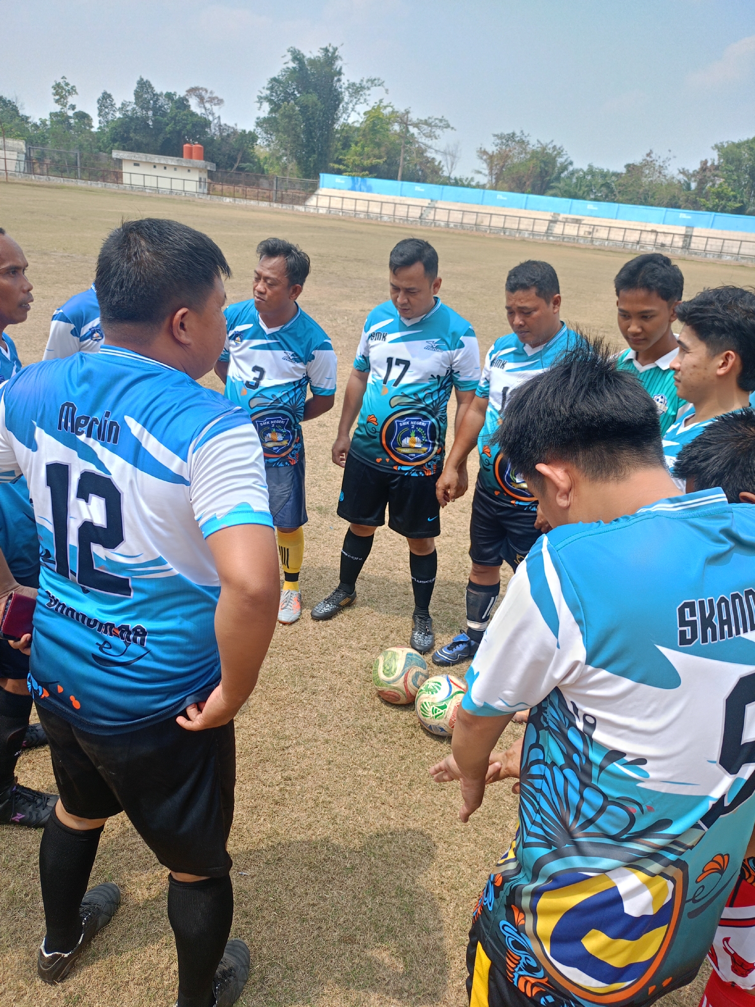 A group of men in blue and white soccer uniforms are gathered in a circle on a dry grass field, seemingly having a discussion. Several soccer balls are on the ground near their feet. In the background, there's a stadium with blue seating and a fence. The sky is clear and bright. The scene appears to be a sports field, likely within a school or community sports complex, given the uniforms which display "SMK NEGERI" (Vocational High School) and the city name "PRABUMULIH". The man on the left of the image is wearing a jersey with the name "Merin" and the number "12" on the back. The overall setting suggests an outdoor sports event or practice taking place in a warm climate.
