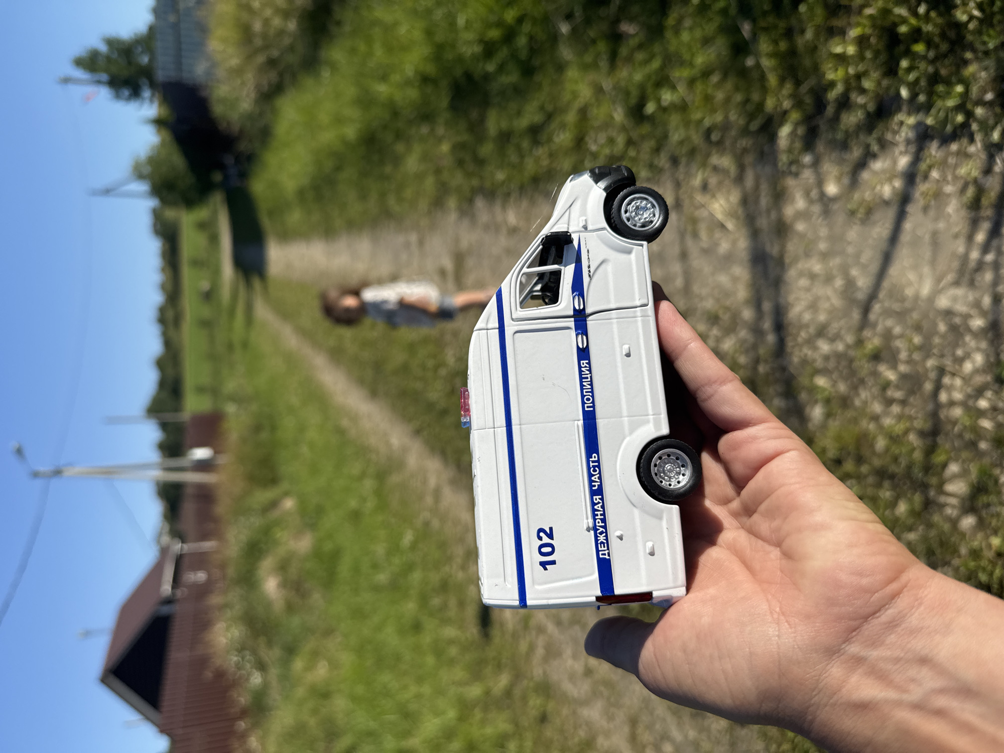 In Kempelevo, Russia, a person's hand holds a white toy police van with blue stripes and the number "102" on its side. The van also has text that reads "DEZHURNAYA CHAST" and "POLITSIYA". In the background, a dirt path stretches through a grassy field, with a blurry figure of a child standing in the distance. Several houses and trees are visible along the sides of the path. The sky is clear and blue.