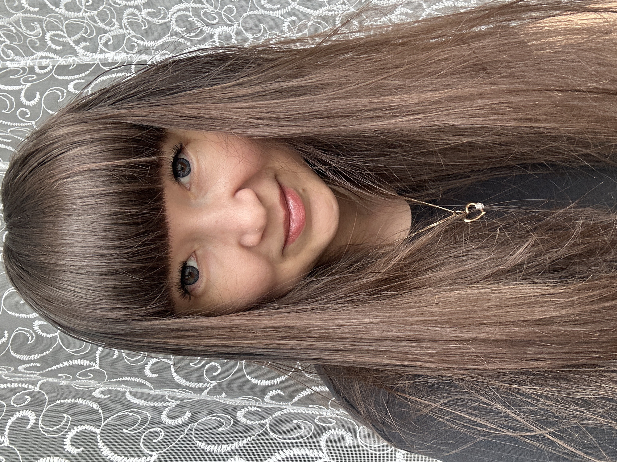 This close-up portrait features a young person, likely taken indoors in Chaikovsky, Russia. The subject has long, straight, medium-brown hair styled with a full fringe (bangs) across their forehead. Their eyes are a striking blue-grey, looking directly at the viewer. A soft, gentle smile graces their lips, which are coated with a light pink lipstick or gloss.
A delicate gold chain necklace with a small, heart-shaped pendant, possibly featuring a tiny clear stone, is visible around their neck. They are wearing a dark-colored top or shirt. The background consists of a patterned, white or light-colored lace-like fabric with swirling embroidery, appearing to be draped over a grey surface, suggesting an indoor setting. The lighting is soft and even, highlighting the subject's features. There is no visible text in the image.