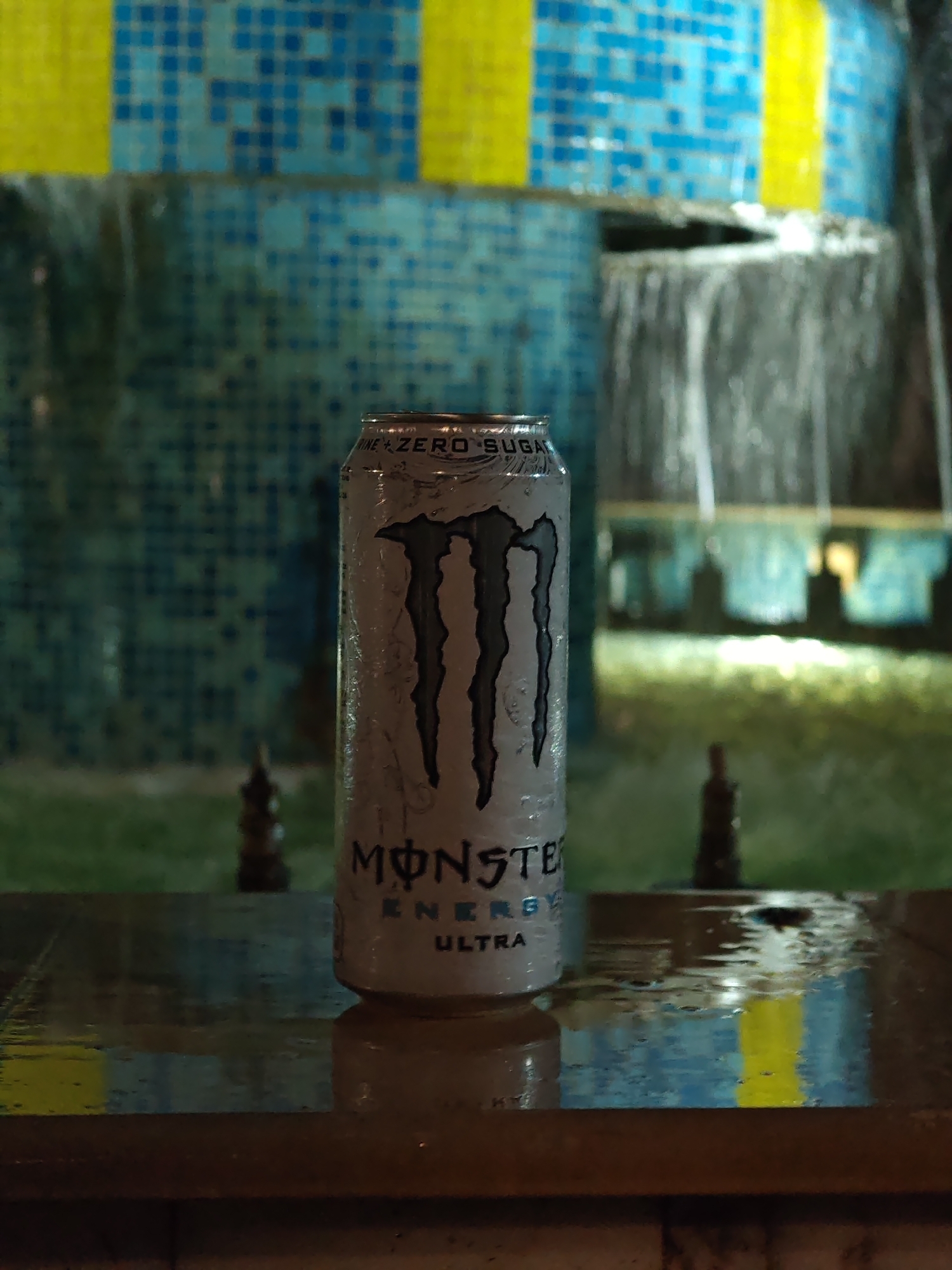 A single, cold Monster Energy Ultra "Zero Sugar" can stands upright on a dark, wet, reflective surface in the foreground. Condensation and water droplets are visible across its white and black metallic body. The surface below reflects the can and elements of the background, also showing numerous water droplets. In the soft-focused background, a tiled water feature or fountain is visible. The tiles are predominantly small squares in various shades of blue, interspersed with sections of yellow tiling. Water cascades downward from a ledge within the structure. The scene is under low, artificial illumination, creating reflections and a distinct atmosphere. No people or other subjects are present. The image is stated to be located in Veliki Preslav, Bulgaria; however, no identifiable landmarks or unique features within the image confirm this specific geographical location.