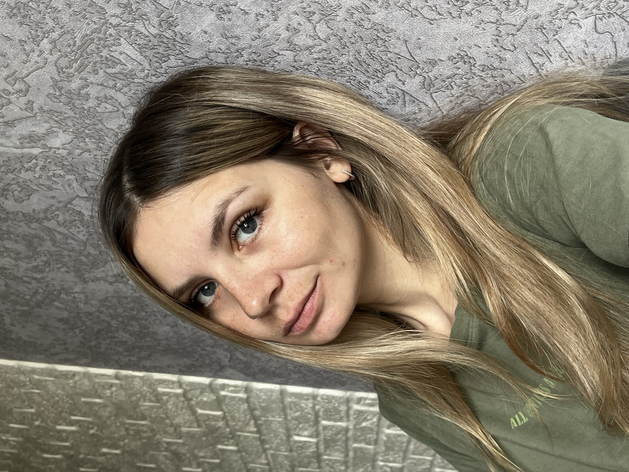 A fair-skinned woman with long, light brown hair poses indoors. She is wearing a green shirt. She has blue eyes. The background includes a textured gray wall and a gray patterned structure. The location is Chaikovsky, Russia.