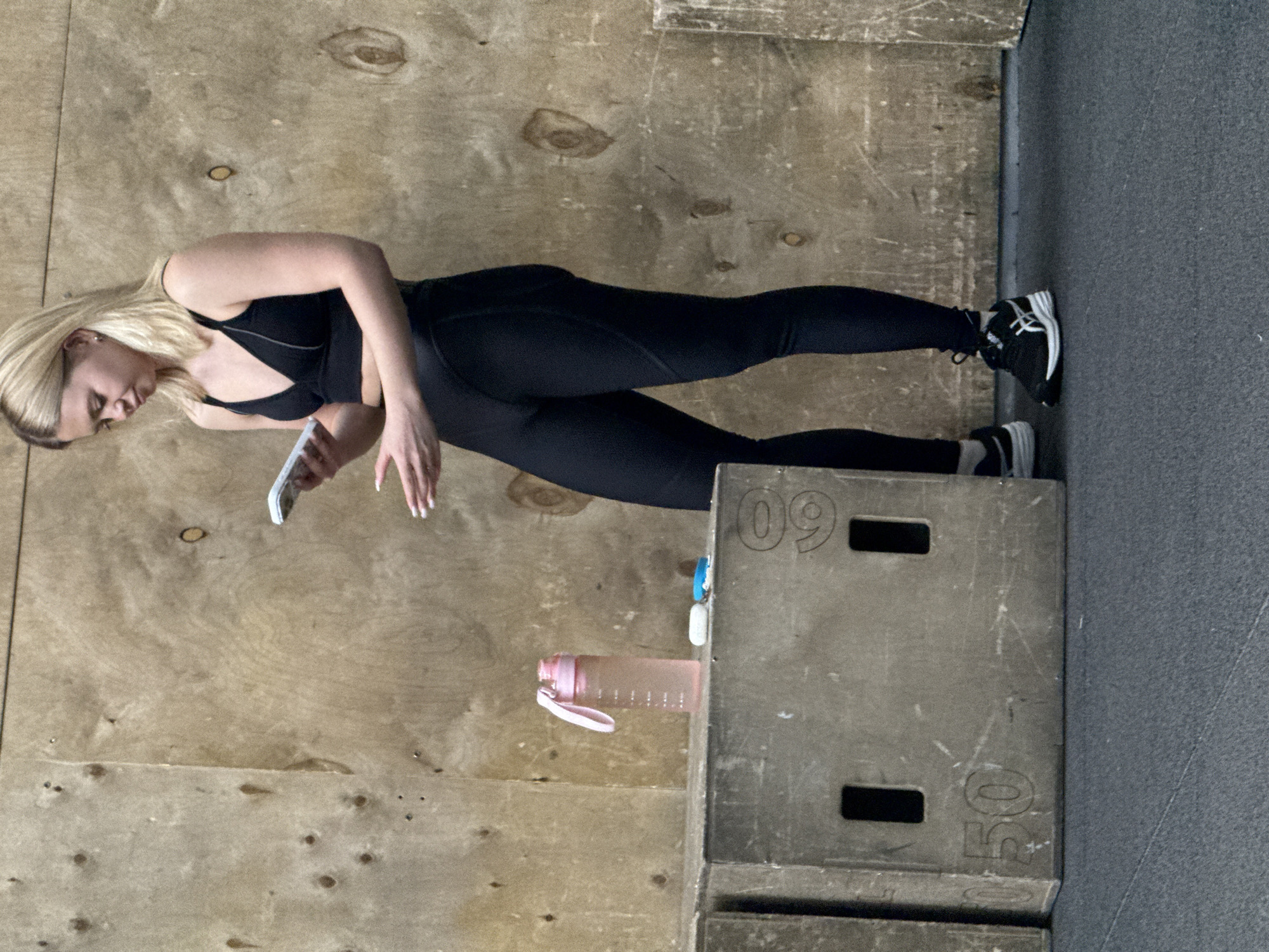 The image, taken in what is contextually identified as Yekaterinburg, Russia, captures a blonde woman in an indoor gym or training facility. She is standing, dressed in black athletic wear consisting of a sports bra with gray trim and full-length leggings, paired with black and white athletic shoes. Her long, straight blonde hair falls around her shoulders as she looks down, focused on a white smartphone held in her left hand. Her right hand hangs loosely by her side. She has light-colored nail polish.
The setting features light-colored wooden paneling on the wall, showing distinct grain and knots. The floor transitions from this wood paneling to a dark gray, textured surface on the right side of the frame. In the mid-ground, a gray, rectangular plyo box or storage box is visible. On its top face, the number "09" is clearly marked, and on its front face, the number "50" is visible. Resting on this box is a translucent pink water bottle with a white lid and strap, alongside a small blue and white plastic clip or charm. The woman appears to be taking a momentary break, possibly checking her phone during a workout.