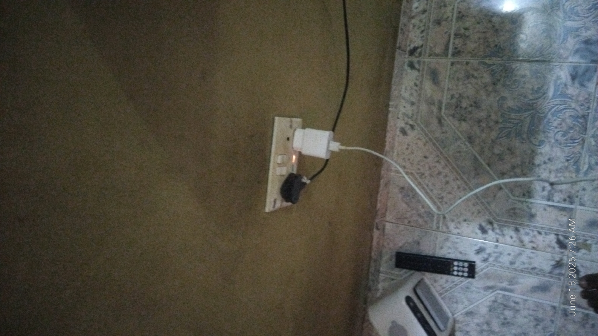 The image shows an electrical outlet on a wall, with two plugs inserted. One plug is black and appears to be a UK-style plug, while the other is a white USB charger adapter. The outlet has two switches, one of which is in the "off" position. There is a small orange light illuminated on the outlet, indicating power.
Below the outlet, on the floor, are a black remote control and a white electronic device with ventilation slots. A white wire runs from the USB charger adapter down to the floor, and another black wire is connected to the UK-style plug. The wall above the outlet is a textured, light brown color. The floor is tiled with a pattern of light and dark grey tiles with a floral design.
A timestamp in the bottom left corner of the image reads "June 15, 2026 7:26 AM." A portion of a person's foot is visible at the bottom of the frame. The scene appears to be an indoor environment, possibly a home or office. There is no indication of the city of Howayi, Nigeria in the image itself.