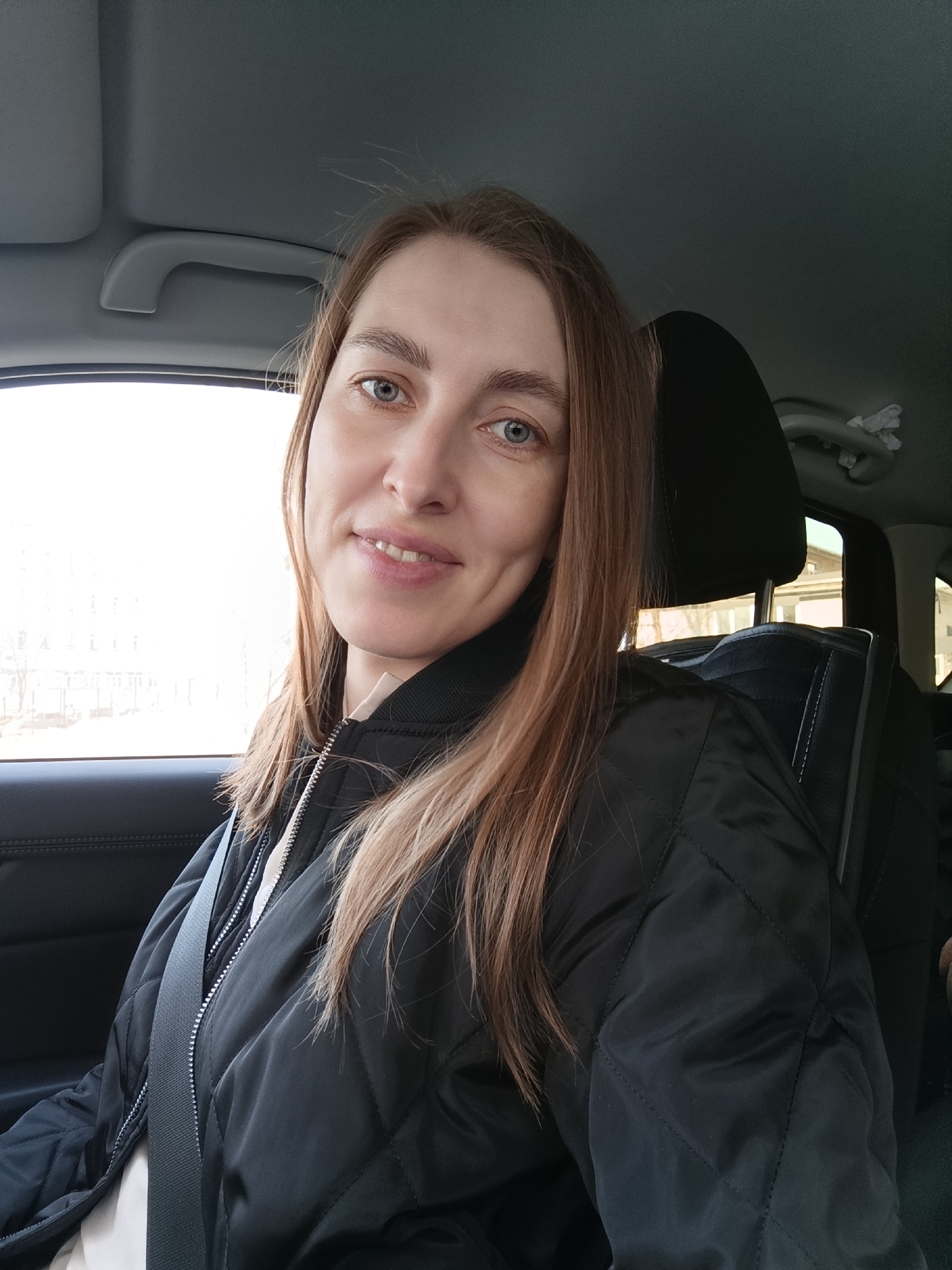 A woman is seated in the driver's seat of a car. She is wearing a black quilted jacket and a seatbelt. The car appears to be in motion, as evidenced by the background visible through the window. Buildings can be seen through the window, suggesting an urban setting in the city of Chaikovsky, Russia. The woman is looking at the camera and smiling. The car's interior includes a headrest, a grab handle, and other components typical of a vehicle.