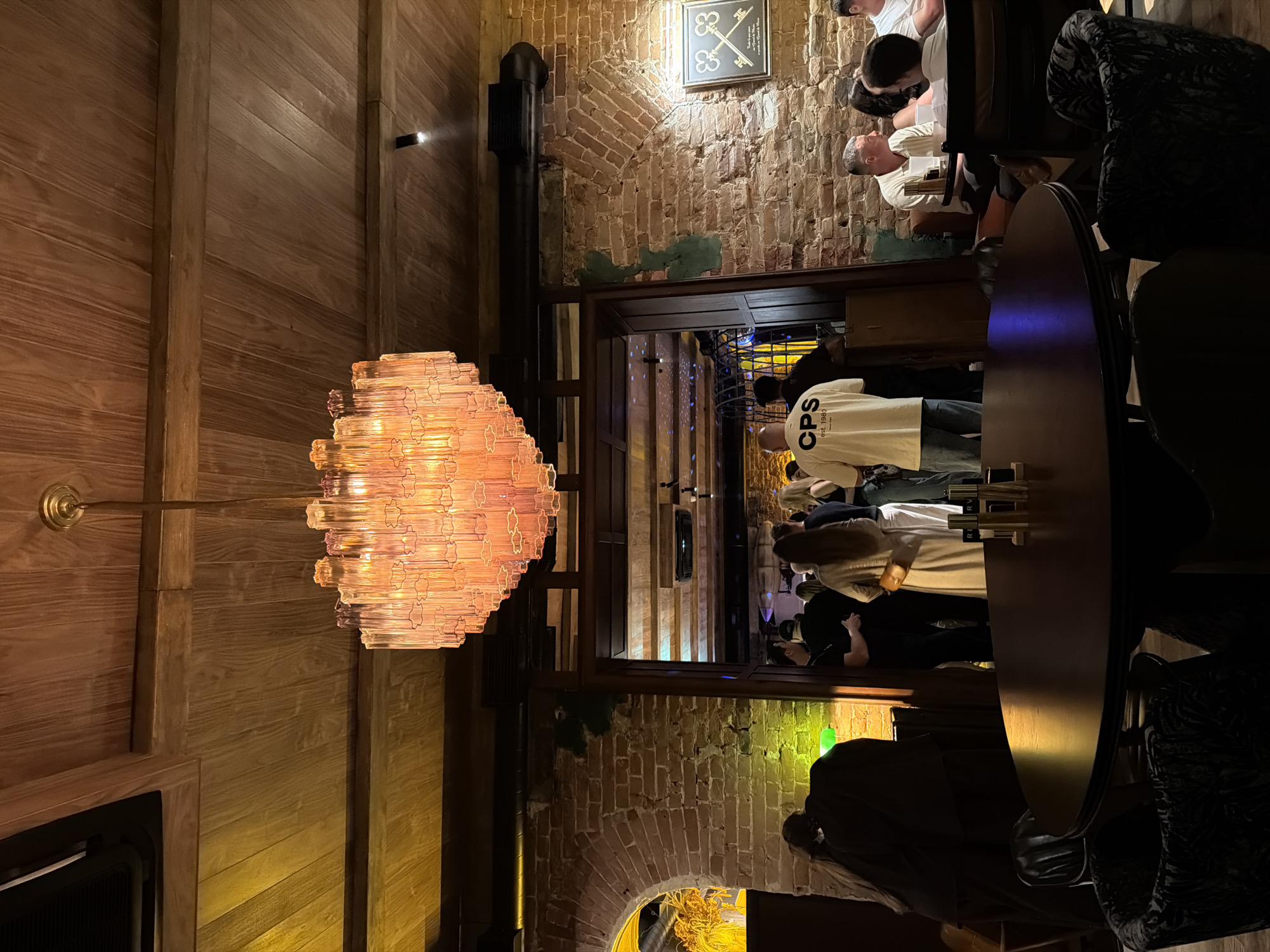 The image depicts the interior of a dimly lit establishment, possibly a restaurant or bar, with a rustic brick wall and a wooden ceiling. A prominent, multi-tiered chandelier made of translucent pink or amber glass hangs from the ceiling. In the foreground, a dark wooden round table is set with what appear to be salt and pepper shakers. Several upholstered chairs with patterned fabric surround the table.
Further into the scene, a doorway or archway leads to another area where a group of people are gathered. Some are standing and conversing, while others are seated at tables. One man in the doorway is wearing a light-colored t-shirt with "CPS est. 1980" printed on it. Another individual is wearing a long, light-colored dress.
To the right, a group of patrons are seated at a table, illuminated by ambient lighting. One man is wearing a white polo shirt and is looking to his left. Another individual is wearing a white t-shirt. A framed picture depicting two ornate keys hangs on the brick wall. The overall atmosphere suggests a social gathering in a stylish, somewhat vintage-themed setting. The context provided about the city of Krasnoyarsk, Russia, is not visually verifiable from the image itself.