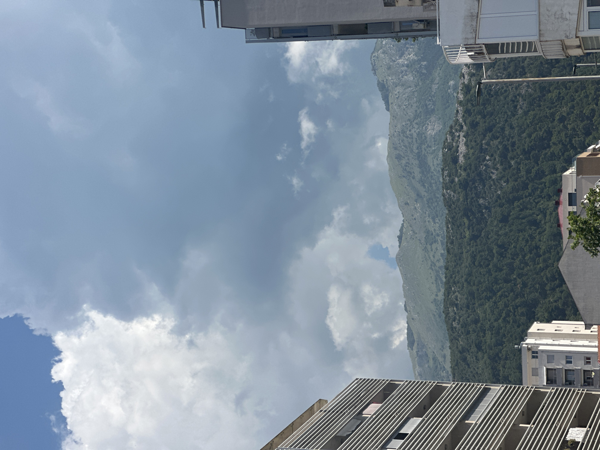 The image captures a view of the mountainous landscape behind the city of Budva, Montenegro. The foreground features modern residential buildings, one on the left with a distinctive ribbed facade and balconies, and another on the right with a white exterior and balconies. In the mid-ground, a dense forest covers the lower slopes of the mountains. The mountains themselves are rugged and rocky, with sparse vegetation visible on their upper reaches. The sky is dramatic, filled with a mix of bright white cumulus clouds and darker, more ominous storm clouds. The overall impression is of a dynamic interplay between urban development, natural beauty, and atmospheric conditions.