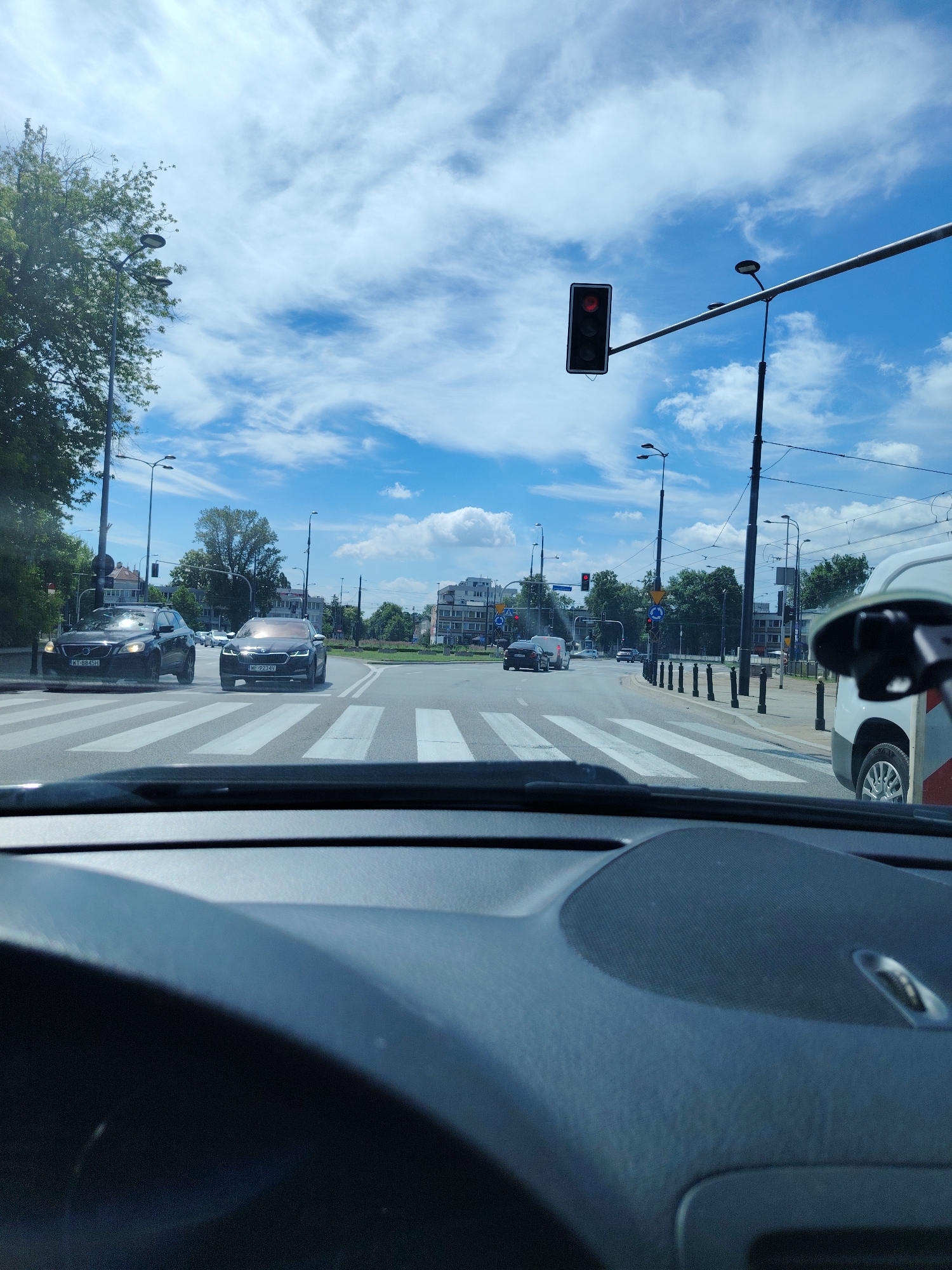 The image, taken from inside a vehicle, shows a street scene in Warsaw, Poland, with a red traffic light visible in the distance. Several cars are stopped at the intersection, waiting for the light to change. A white van is parked on the right side of the road. The sky is blue with scattered white clouds. The intersection features a pedestrian crossing and several streetlights. Buildings and trees line the sides of the road. The interior of the vehicle, including the dashboard and steering wheel, is visible in the foreground.