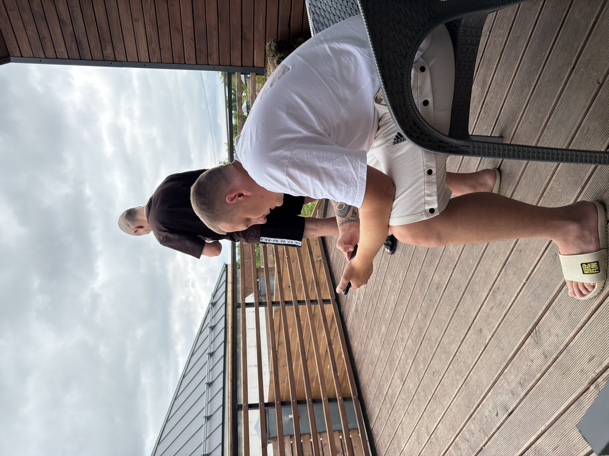 Two men are on a wooden deck. One man is standing and looking out. The other man is sitting in a chair and looking at his phone. The deck has a wooden railing and a wooden roof. The sky is cloudy. The scene is in the city of Semiluki, Russia.