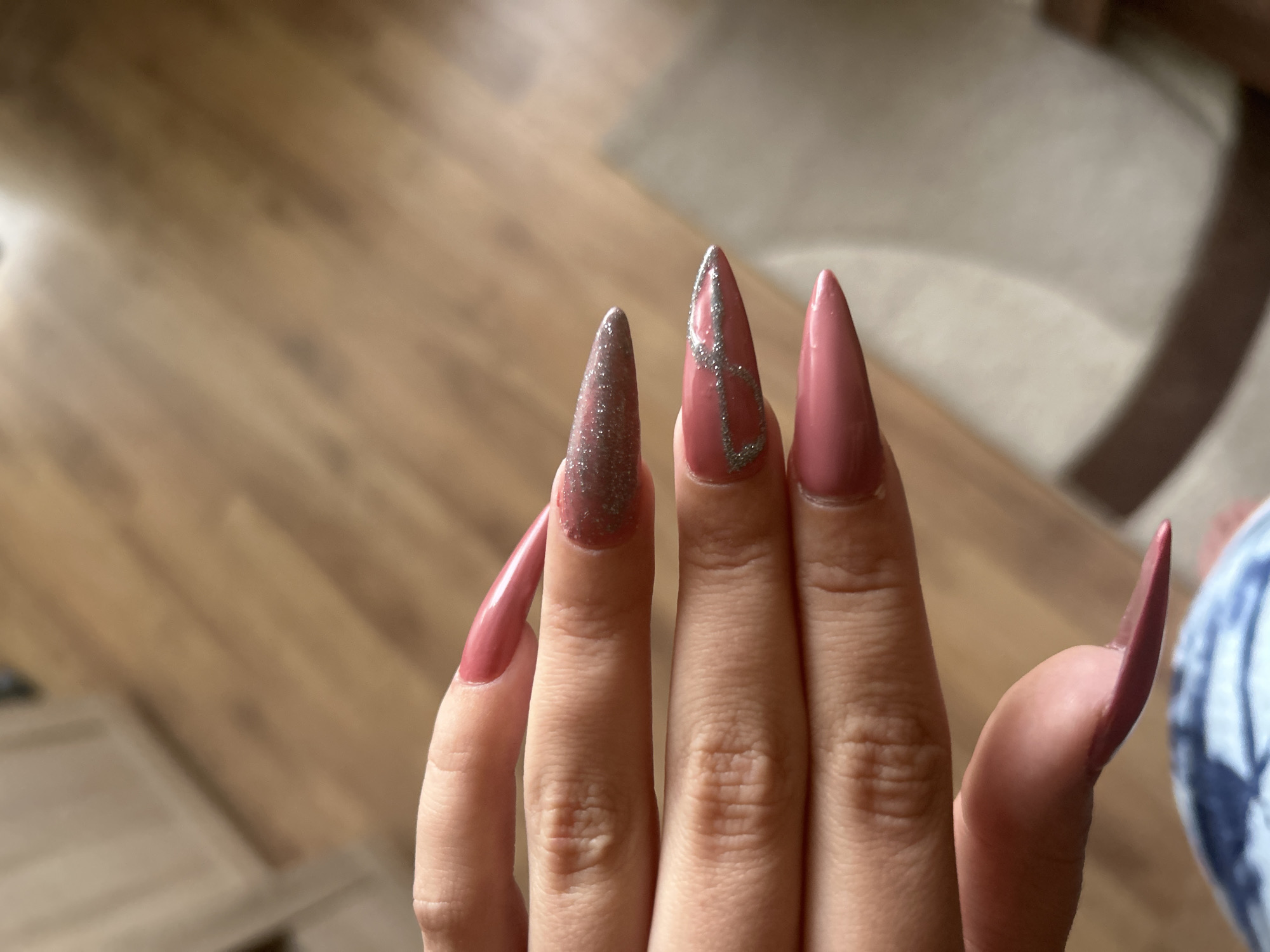 A close-up shot showcases a hand displaying a set of long, stiletto-shaped nails. The nails are painted in a mauve color, with the index finger nail featuring a sparkling silver ombre effect from the base to the tip. The middle finger nail is adorned with a silver glitter outline design resembling a stylized number 8 or infinity symbol. The background consists of a blurred interior scene, including wooden flooring and a glimpse of a light-colored rug. The scene appears to be indoors. No city or location is discernable.