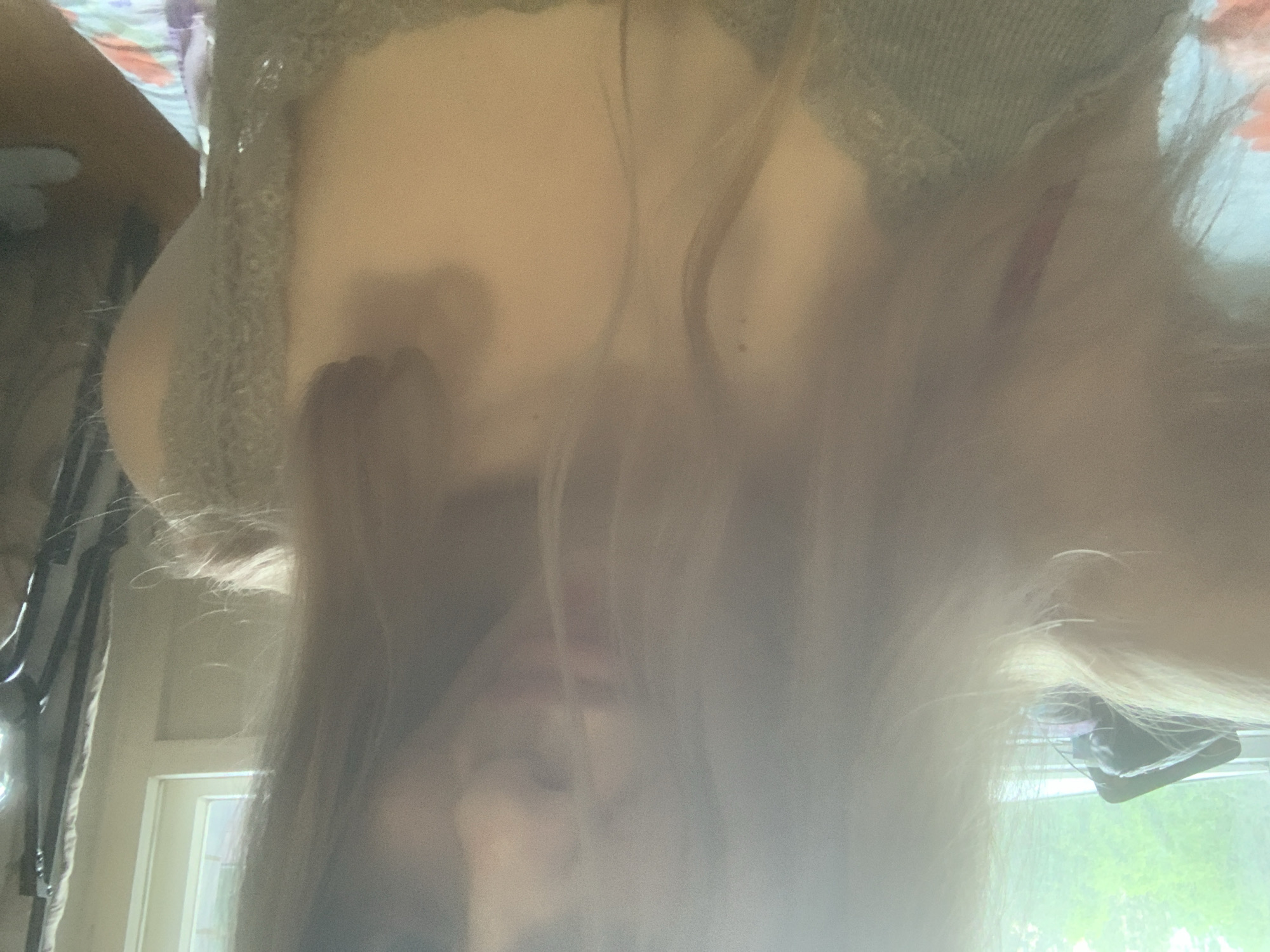 The media file is a brightly lit, upside-down selfie featuring a person, likely female, with long, light-colored hair. The individual is positioned looking downwards at the camera, causing their hair to fall towards the top of the frame, obscuring much of their face due to the angle, strong light, and blur.
The person appears to be wearing a light-colored garment, possibly a sweater or shawl, with a textured, knitted, or crocheted pattern.
The scene is set indoors during the daytime, indicated by the abundant natural light streaming in from a window in the background. Green foliage is visible outside the window, suggesting a warm season. Hints of an interior space include a door or door frame and a dark, possibly fabric object or bag hanging near the window.
The primary activity is the act of taking an upside-down selfie. Notable details include the strong light source, the blurry and somewhat overexposed quality of the image, and the unique perspective. No text is visible in the image. The location context is Unknown, Russia.
