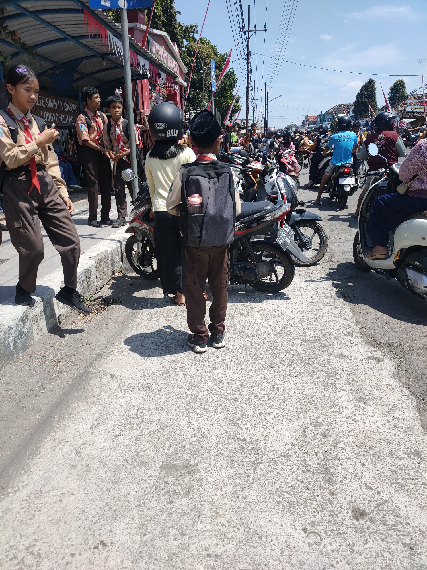 The image depicts an outdoor scene in Kanigoro, Indonesia, on a sunny day, with a street lined by a building adorned with red and white flags and banners. Several individuals are present, many of them appearing to be students in school uniforms consisting of tan shirts and brown pants, some wearing red neckerchiefs and scout badges. A young girl in the foreground, on the left, is wearing a tan uniform and a red neckerchief, holding a mobile phone. Beside her are two young males in similar uniforms, one holding a red and white flag. A young boy in a tan uniform and a black skullcap, carrying a gray backpack with a pink water bottle holder, stands in the middle of the street. Another student in a tan uniform and black helmet is also visible.
The street is filled with motorcycles and scooters, suggesting a busy event or gathering. Vehicles are parked and some individuals are seated on them. The road surface is made of concrete. In the background, more people and vehicles are visible, along with a street with buildings and trees. The sky is clear and blue. The overall atmosphere appears to be one of a community event or a school activity taking place on a public street.