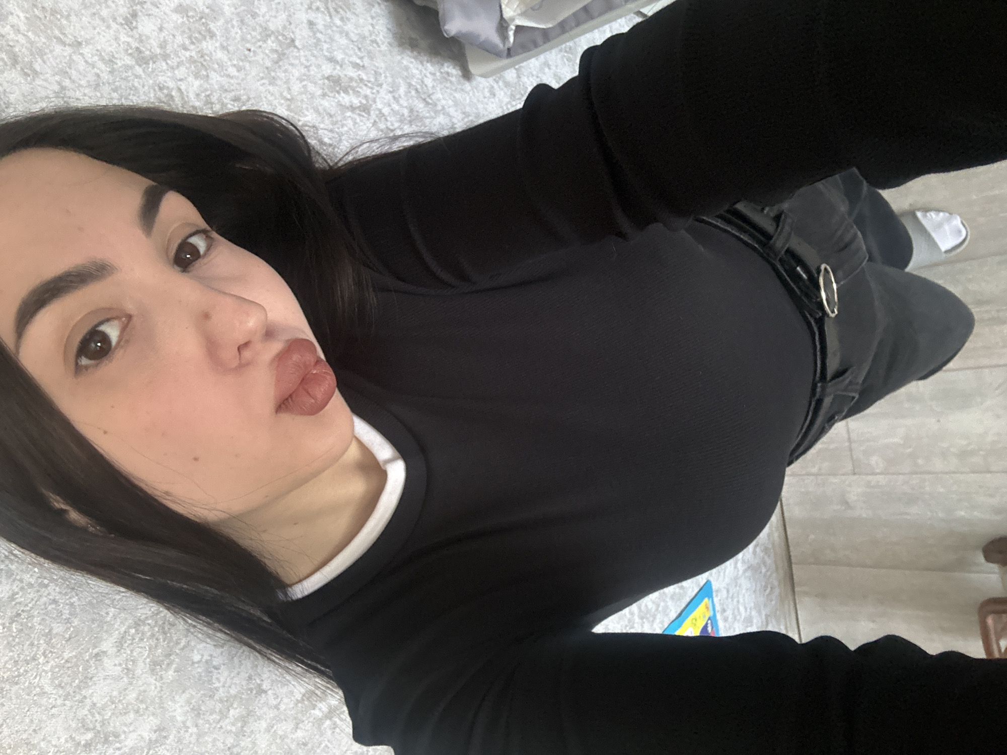 A woman lies on a light gray textured surface that appears to be a carpet or rug, likely indoors. She is dressed in a black long-sleeved shirt with a white collar, black pants with a black belt, and white slippers are partly visible. Her dark hair is loose around her face. The background also includes a floor and some household items. It is not possible to verify the location in the city of Марковский, Russia, or to infer any information about specific actions or interactions.