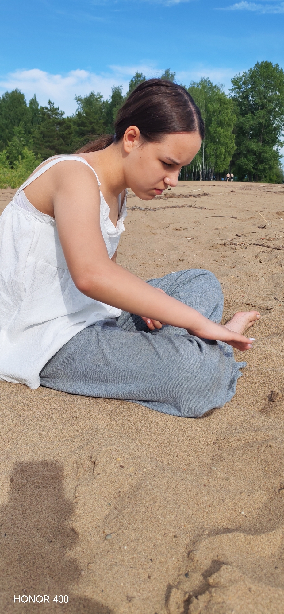An outdoor, eye-level shot captures a young female sitting on a sandy beach on a sunny day. The subject, likely a teenager with dark hair pulled back in a ponytail, is wearing a white sleeveless top and light grey pants. She is seated with her legs bent, her right hand extended towards the sand, seemingly playing with or drawing in it. Her gaze is directed downwards, focused on her activity.
The setting is a wide expanse of light brown sand. In the background, a dense line of green trees, including both conifers and deciduous types, stretches across the horizon. Above the trees, a bright blue sky with scattered white clouds indicates clear weather. The strong sunlight casts distinct shadows on the sand, including a partial shadow of the photographer in the bottom left. Two small, indistinct figures, presumably people, can be seen further down the beach near the tree line in the distance. The overall atmosphere is one of calm engagement with nature. In the bottom left corner of the original image, the text "HONOR 400" is visible, indicating the device used to capture the photo.