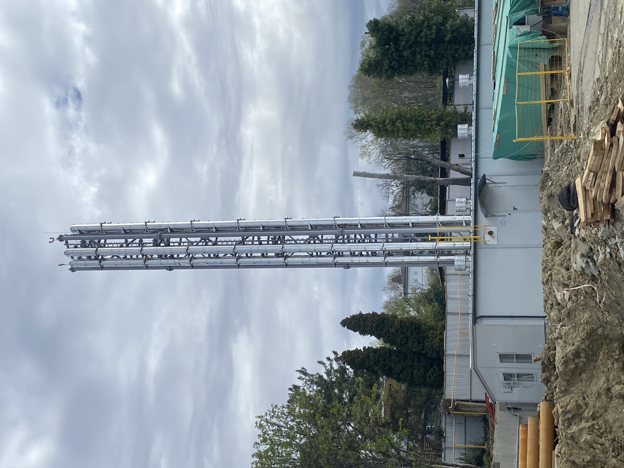 The image depicts an industrial scene likely located in or near Sochi, Russia. Dominating the left side of the frame is a tall, engineered structure composed of multiple parallel pipes supported by a network of metal beams. These pipes extend upwards, seemingly connecting to a facility obscured from full view. To the right of the piping structure is a light-colored, single-story building with at least two visible windows. Yellow safety fencing and green tarpaulin cover sections of the building's exterior. Adjacent to the building, on the right, is a pile of dirt and lumber, suggesting construction or renovation activities. Trees line the mid-ground, adding a touch of natural environment to the otherwise industrial landscape. The sky is overcast with grey clouds, indicating a cloudy day. There are no visible people in the frame. Based on the construction style and infrastructure, this could be an energy plant, a water treatment facility, or similar industrial site.