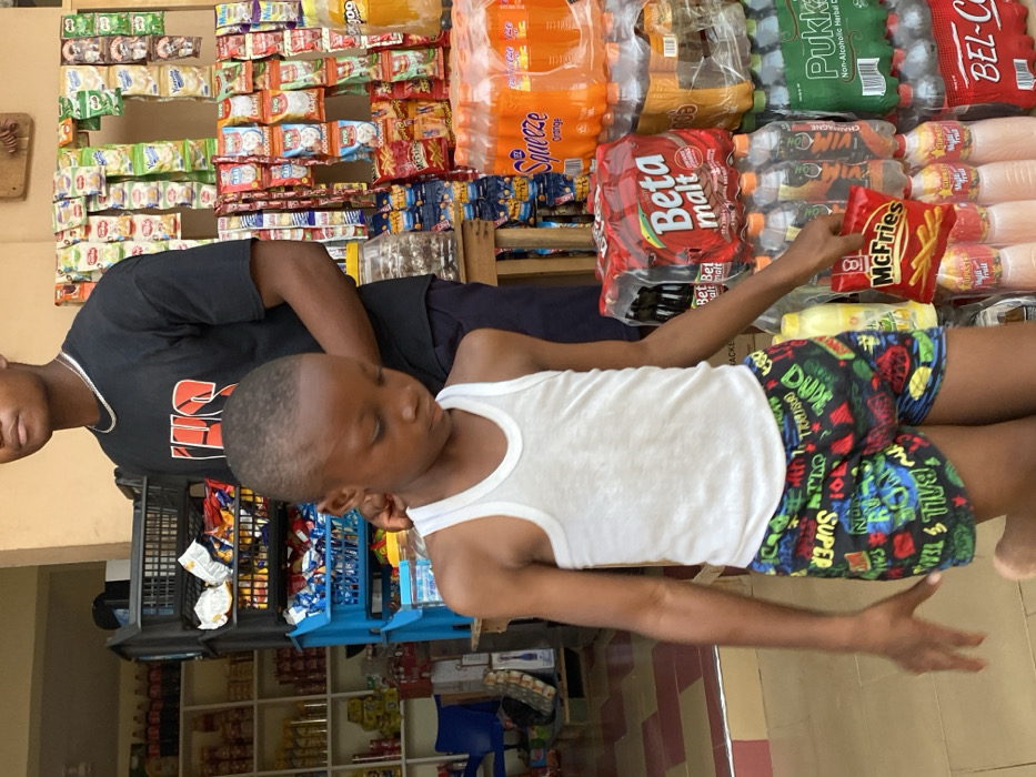 An outdoor market scene presents two people. An adult with a black t-shirt stands near a boy wearing a white tank top and colorful cartoon-printed shorts. The boy holds a bag of "McFries." The market behind them is stocked with various items: beverages like "Squeeze Orange" and "Beta Malt," alongside packaged snacks and drinks. The merchandise is neatly arranged on shelves, creating a vibrant backdrop. The scene suggests a casual moment captured amidst a local commercial setting.