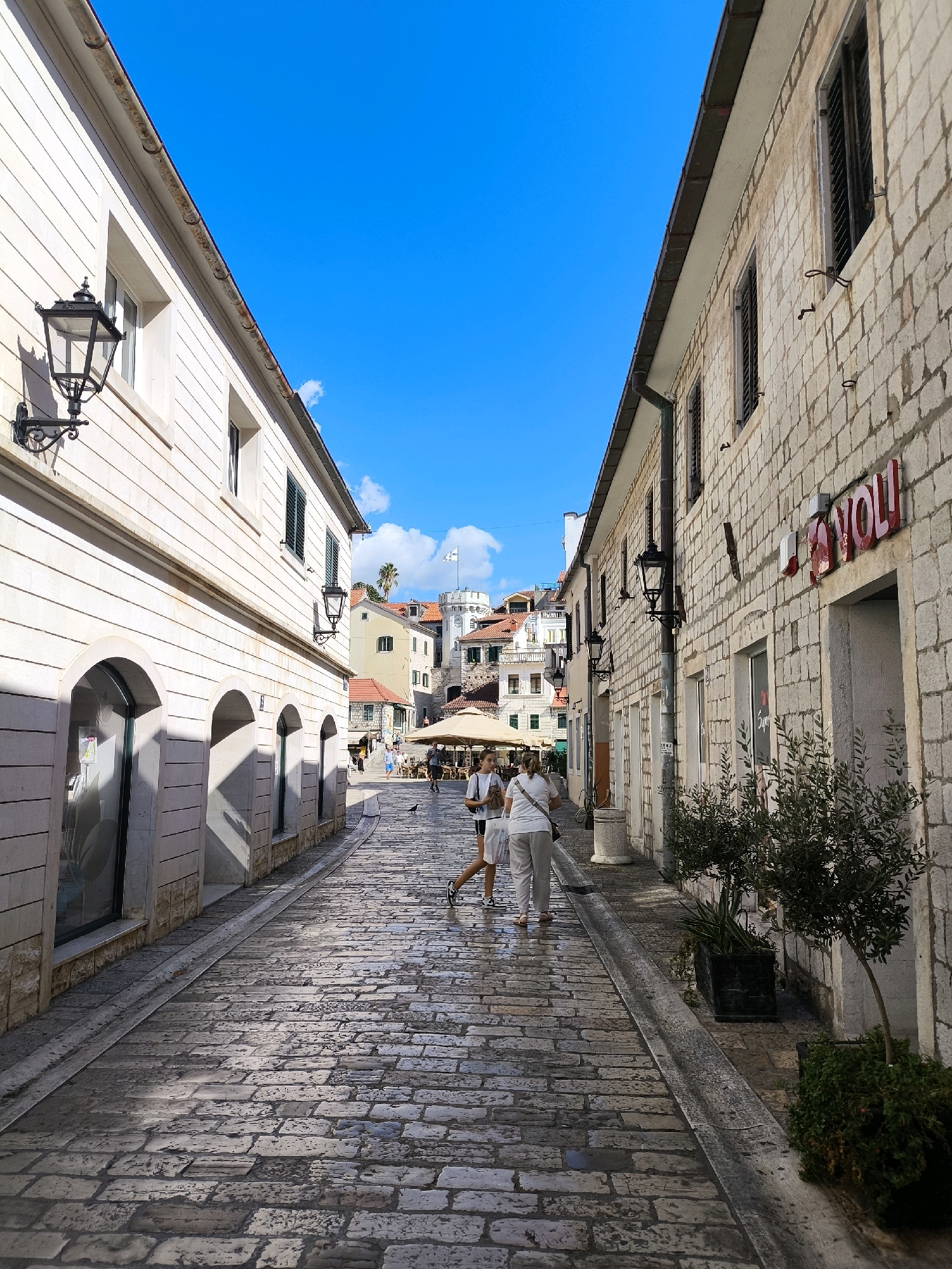 The image shows a narrow, cobblestone street in a European city, likely Herceg Novi, Montenegro, given the architectural style and vegetation. The street is flanked by two buildings with stone facades. The building on the left has a pale stone finish with arched storefronts and traditional black lanterns mounted on the wall. The building on the right is constructed of lighter-colored rough-hewn stone blocks and features shuttered windows and a red neon sign with the word "JOLI" visible. Several black, ornate lanterns are mounted on poles along this side of the street.
In the background, more buildings with red-tiled roofs are visible, suggesting a densely populated urban area. A tower with a flag is prominent in the distance. Two women are walking away from the camera down the street. One woman is wearing a white t-shirt and grey pants, and the other is in a white top and shorts. They are walking on the wet cobblestone street, which reflects the bright blue sky. There are potted plants and trees on the right side of the street. The overall impression is a sunny day in an old, charming European town.