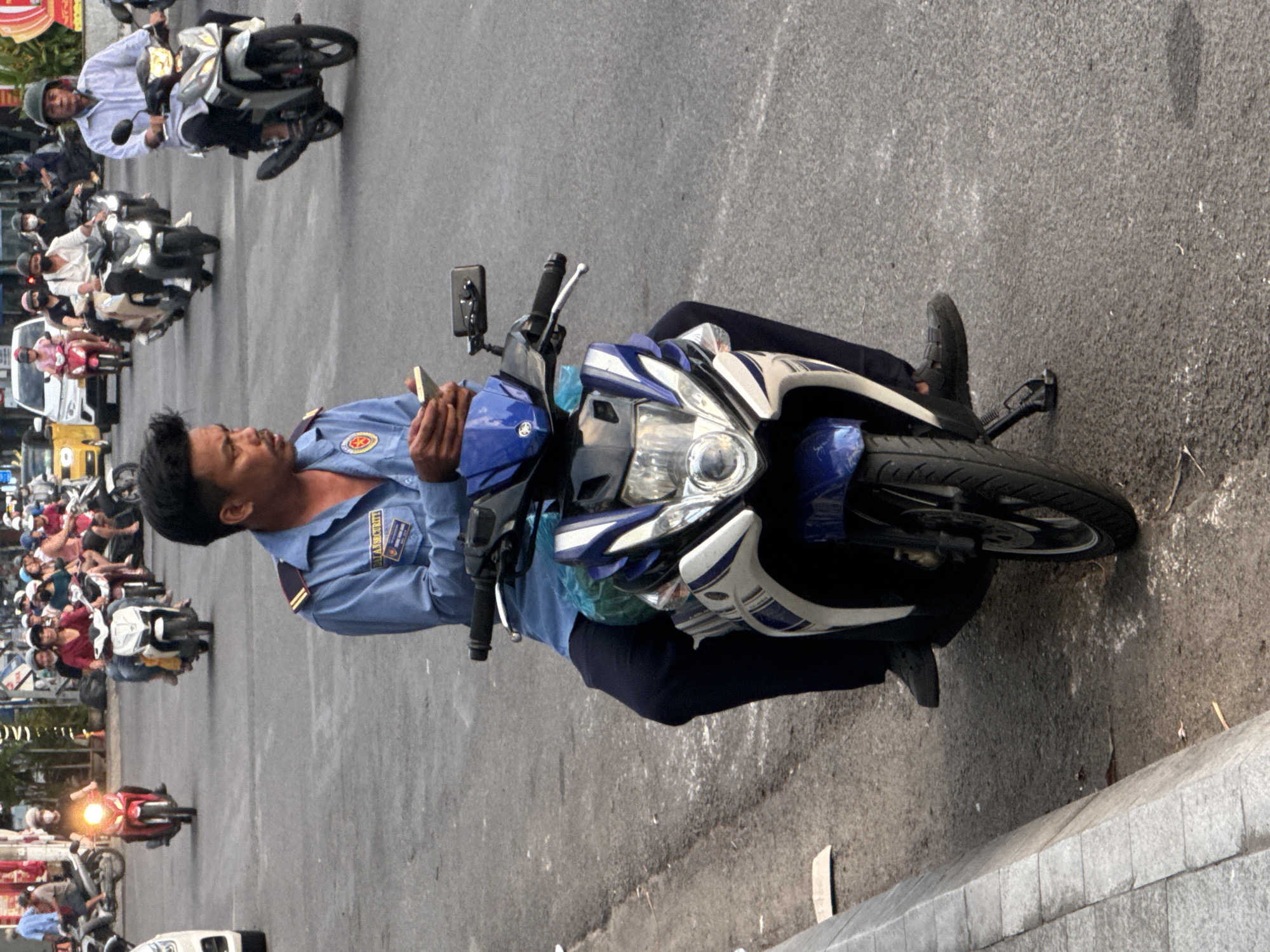 In Nha Trang, Vietnam, a man wearing a blue uniform with a yellow and red emblem is sitting on a blue and white motorcycle. He is holding a smartphone in his hands, looking down at it. The motorcycle has a black seat and handlebars. The man is wearing dark pants and dark shoes. In the background, other motorcycles and cars are visible on a paved road. Some of the vehicles are occupied by people. The scene appears to be on a city street with buildings and foliage lining the sides of the road. The lighting suggests it is daytime.