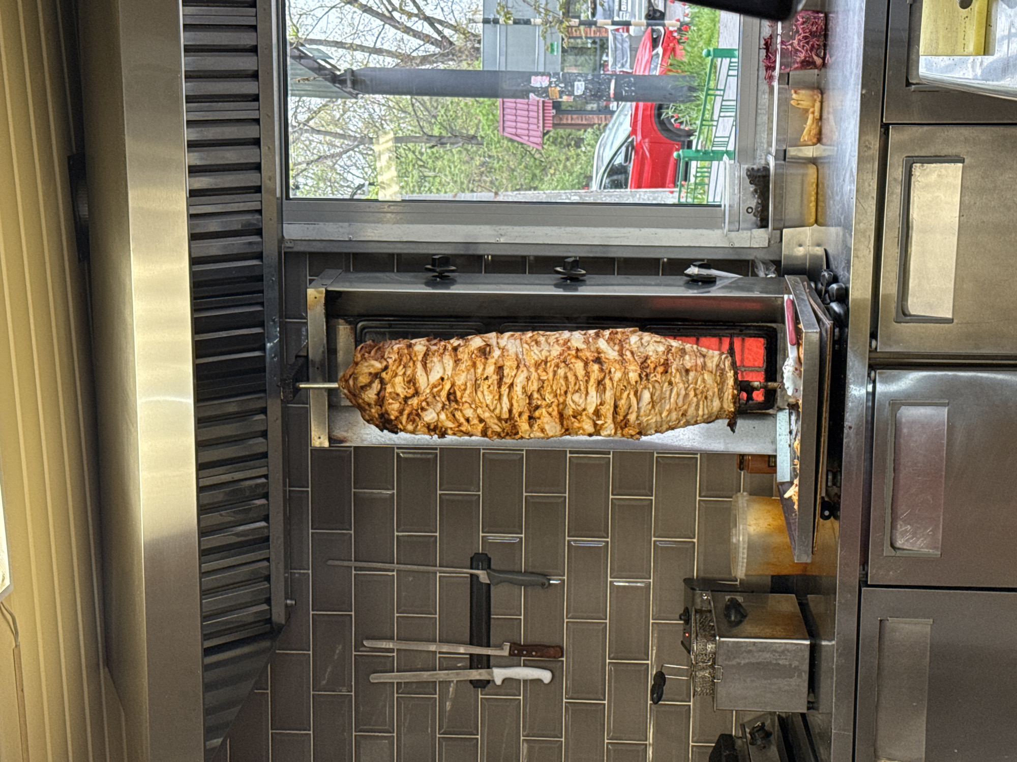 The image captures an interior view of a commercial food preparation area, likely a shawarma or doner kebab stand, focusing on the cooking process. A large, cylindrical skewer of seasoned meat, predominantly light brown and textured, is rotating on a vertical rotisserie. Red-hot heating elements are visible on the right side of the meat, indicating active cooking.
Below the rotisserie, a gray tiled wall serves as a backsplash, on which three knives with different handles (one black, one wooden, one white) are neatly arranged, possibly held by a magnetic strip. To the far left, a light-colored, ribbed panel is visible, contrasting with the metallic surfaces of the kitchen equipment.
To the right of the rotisserie, there are various stainless steel kitchen appliances, including what appears to be a smaller countertop unit with knobs and switches. Above this area, near a window, several clear plastic containers hold ingredients; one contains finely chopped red ingredients (possibly cabbage or sumac onions), and another contains yellow ingredients (perhaps corn or chickpeas).
Through the window in the upper half of the image, an outdoor street scene is visible. Green trees with sparse foliage suggest late spring or early summer. A light pole or sign is prominently featured, and a red car is parked or driving on what appears to be a street or parking lot. The overall lighting suggests it is daytime. No people are visible in the frame. The scene conveys a functional and busy food service environment in Rostov-on-Don, Russia. No discernible text is visible on any objects.
