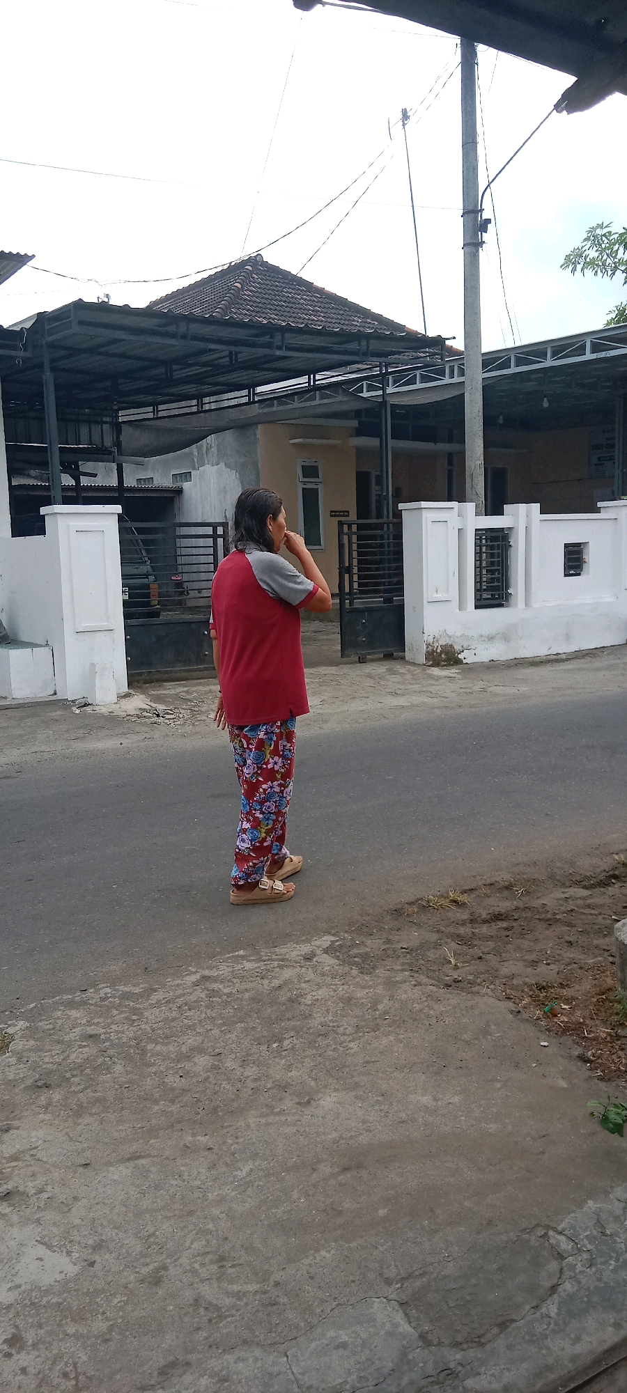 A woman stands on a paved road, facing away from the camera and to the right. She is wearing a red t-shirt with gray sleeves and red pants with a floral pattern. She has on tan sandals. Her right hand is near her mouth, and her left hand is by her side. Behind her are several buildings with covered carports, fences, and a white wall with decorative elements. A utility pole with wires runs behind the buildings. The sky is overcast. The scene appears to be in a residential area.