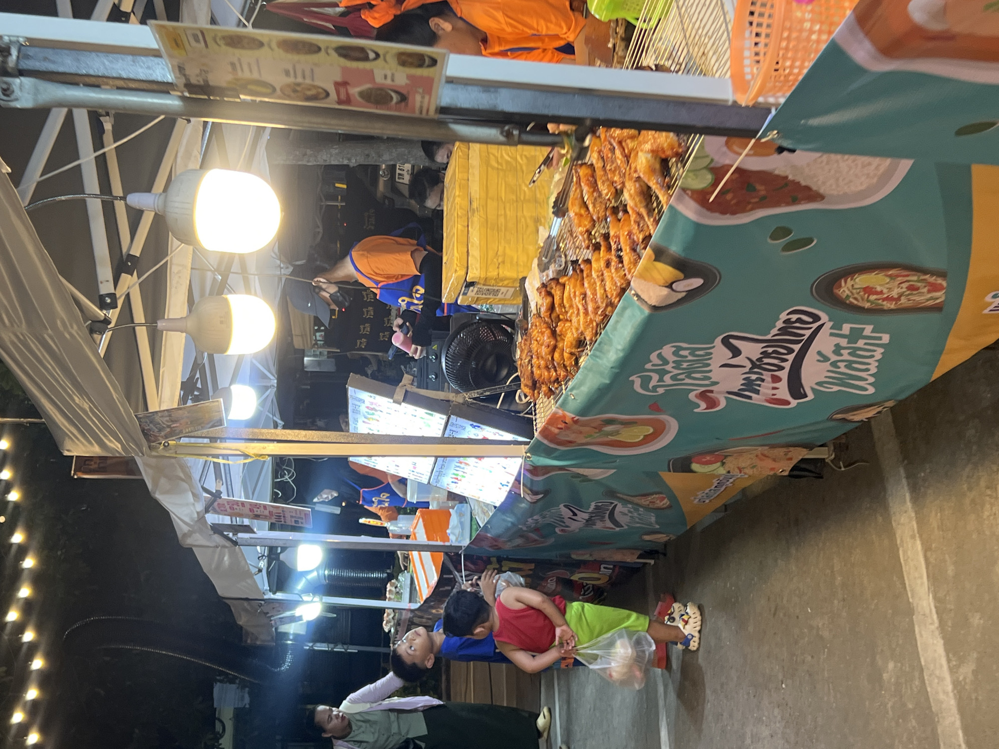 A night-time outdoor food stall scene in Choeng Thale, Thailand, features a row of grilled food, likely chicken, on display under bright lights. Two young boys are looking at the food, one in a red shirt and green shorts holding a plastic bag, and the other in a blue shirt. A woman in a light-colored shirt stands to the left, appearing to be an adult accompanying the children. Vendors in orange shirts are visible behind the stall, serving customers. A green banner with images of food items and Thai text is draped along the front of the stall. The ground is paved, and string lights illuminate the area.
