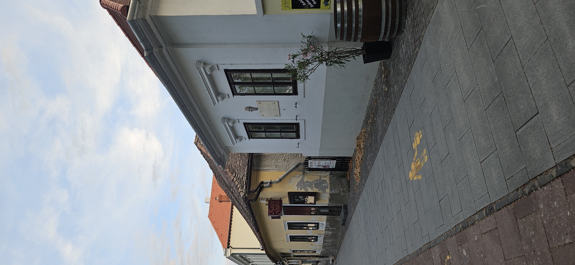The image shows a street scene in Keszthely, Hungary, likely in the historic town center. A pedestrian sidewalk is paved with large gray rectangular tiles. On the sidewalk, a yellow stencil depicts two figures, one larger and one smaller, connected by a hand, possibly indicating a family or child-friendly path. Further down the sidewalk, another yellow stencil of an arrow points to the left.
Two buildings line the right side of the street. The building in the foreground is a white, one-story structure with a gray lower facade. It has dark-framed windows and decorative white trim above them. A plaque with a relief of a human head and text is mounted on the wall between two windows. A potted plant with pink flowers stands to the right of the windows, and a wooden barrel, likely decorative, is placed next to it.
Behind the white building, a yellow building is partially visible. It has a red-tiled roof and a sign above its entrance that reads "Kastély Galéria." The yellow building appears older and has peeling paint in some areas. A dark wooden gate with a poster attached is located to the right of the yellow building's entrance.
The street slopes gently downward from left to right. The sky above is partly cloudy with a mix of blue sky and white clouds. Fallen leaves are scattered on the ground near the buildings. The overall impression is of a quiet, historic European street.