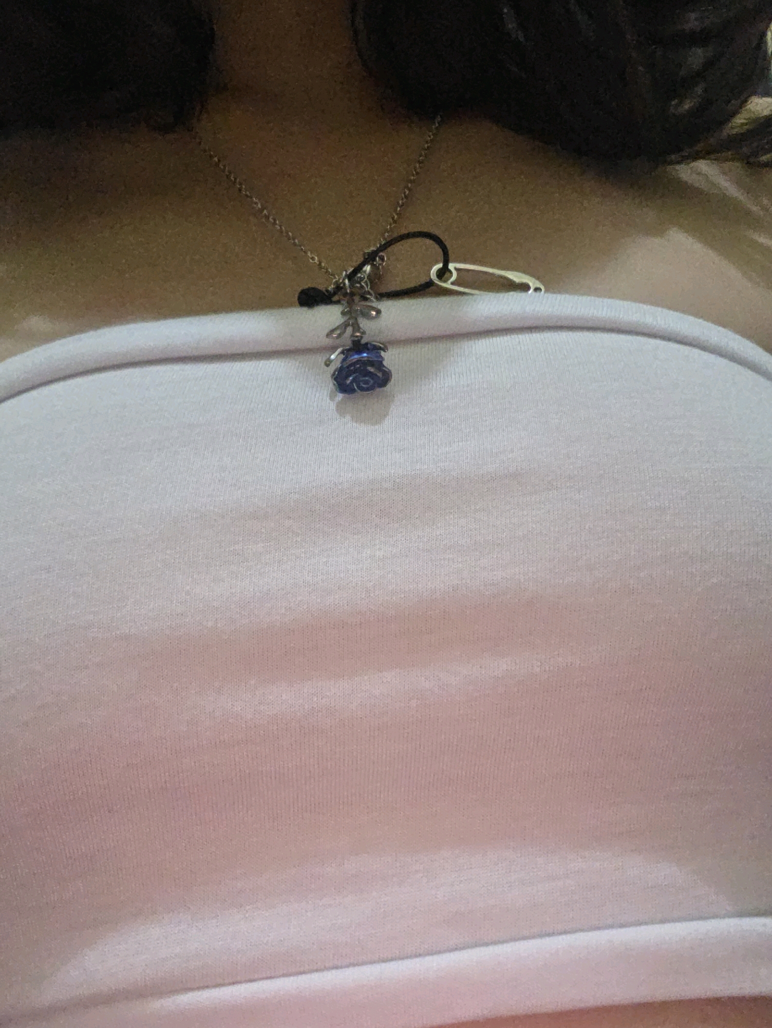 The image shows a close-up view of a person wearing a light pink strapless top. A silver chain necklace with a dark cord and a small blue rose pendant is visible on the person's chest. A safety pin is attached to the dark cord of the necklace. The background is dark and out of focus, suggesting the photo was taken indoors or in low light conditions. There are no discernible landmarks or features that would indicate a specific location in Phan Thiết, Vietnam.