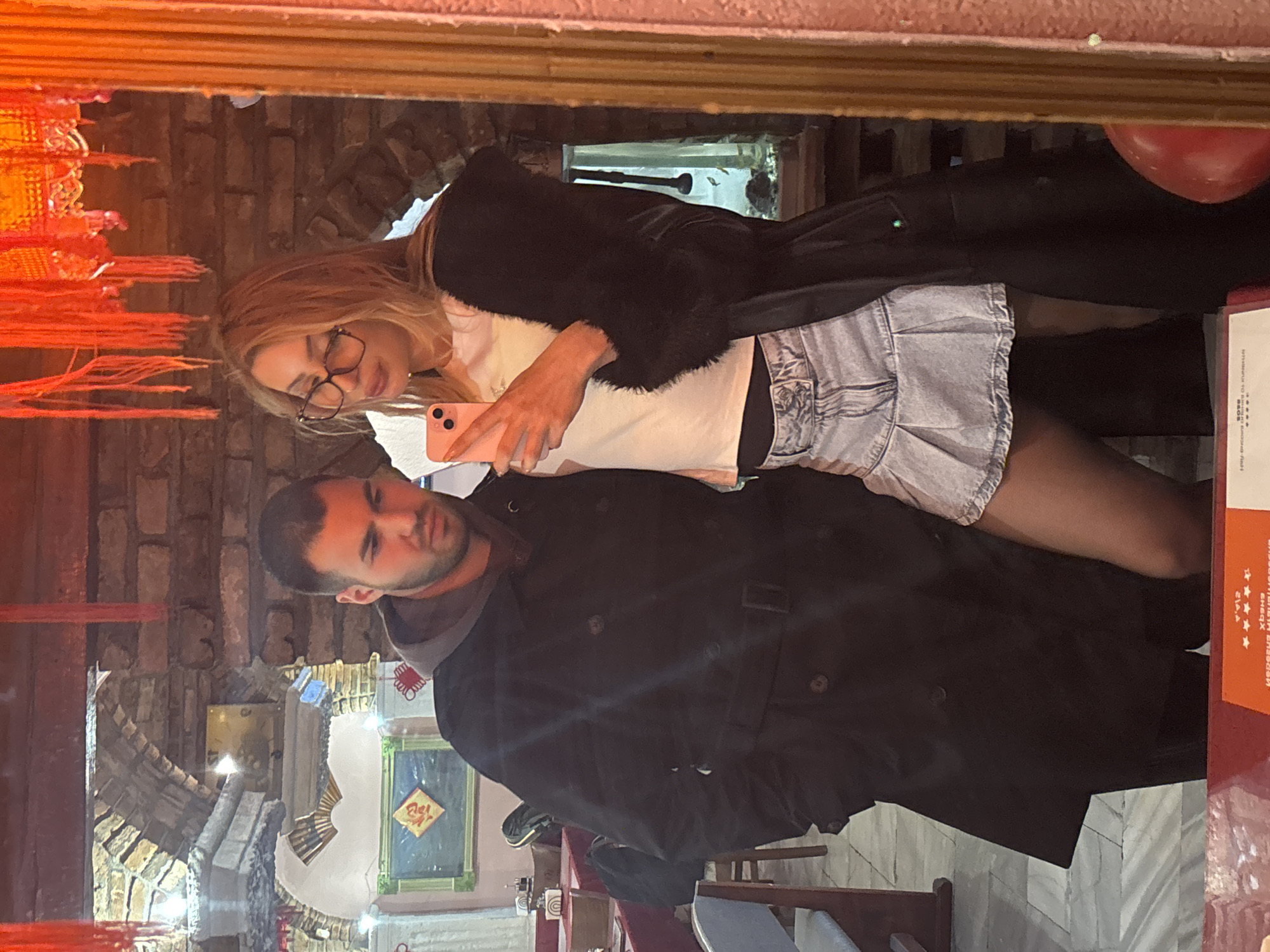 The image shows a reflection in a mirror, depicting a couple in an indoor setting. The woman has blonde hair, wears glasses, and a light-colored top with a denim skirt under a black coat with fur trim. She holds a pink smartphone. The man has dark hair and wears a black coat. The mirror reflects a room with decorative elements, including a brick wall, and decorative fixtures. Behind the people are chairs and tables. Given the image context and architecture, the location is consistent with a restaurant interior in Varna, Bulgaria.