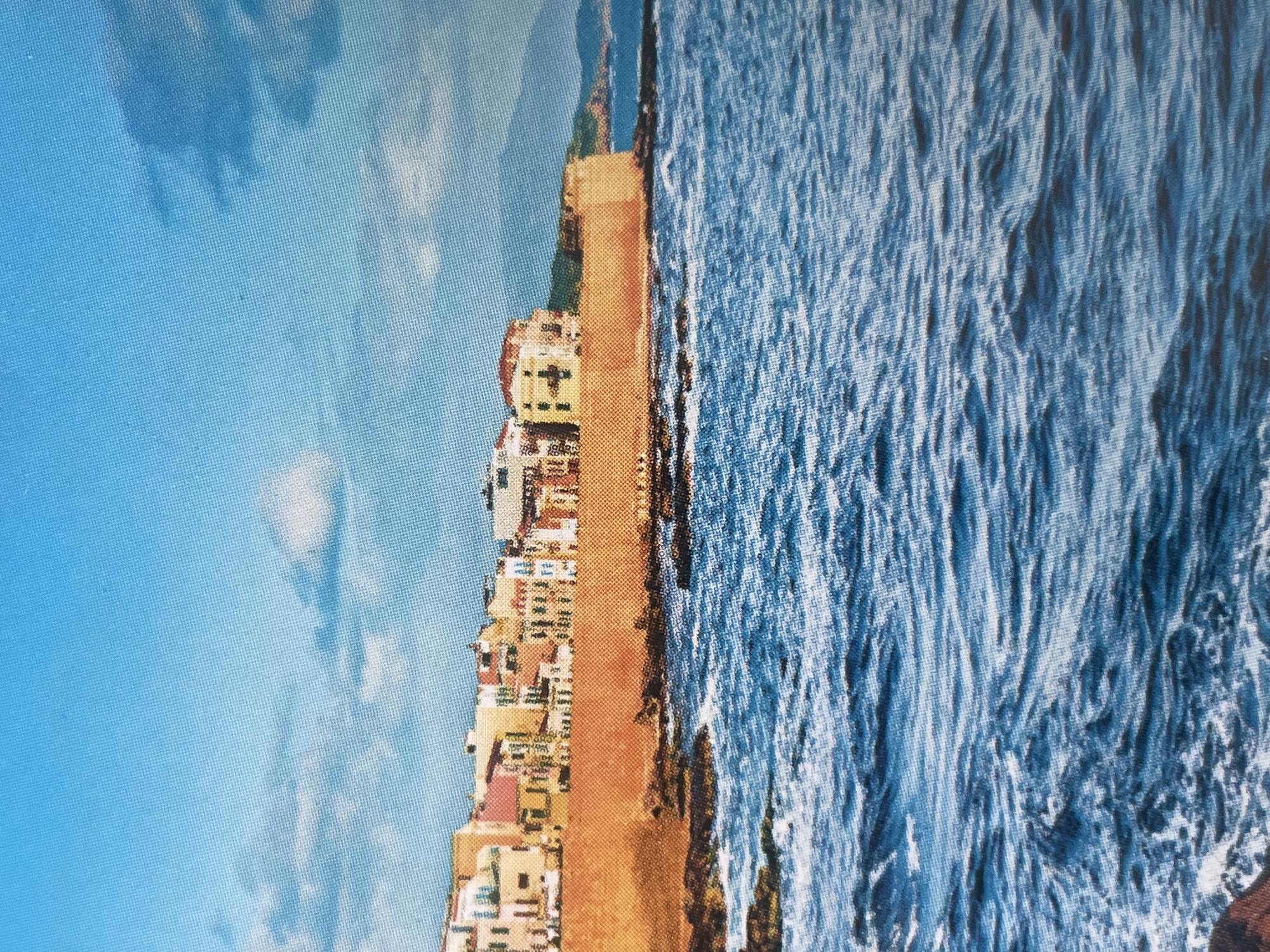 The image depicts a coastal city with colorful buildings situated behind a large, orange-hued wall that appears to be a fortification. The buildings have a Mediterranean architectural style, with various shades of yellow, pink, and white. In front of the wall, there are rocky outcrops. The sea in the foreground is a deep blue with visible waves and foam. The sky above is a bright blue with scattered white clouds. Distant hills are visible on the horizon. There are no people or any indication of the city being Sofia, Bulgaria.