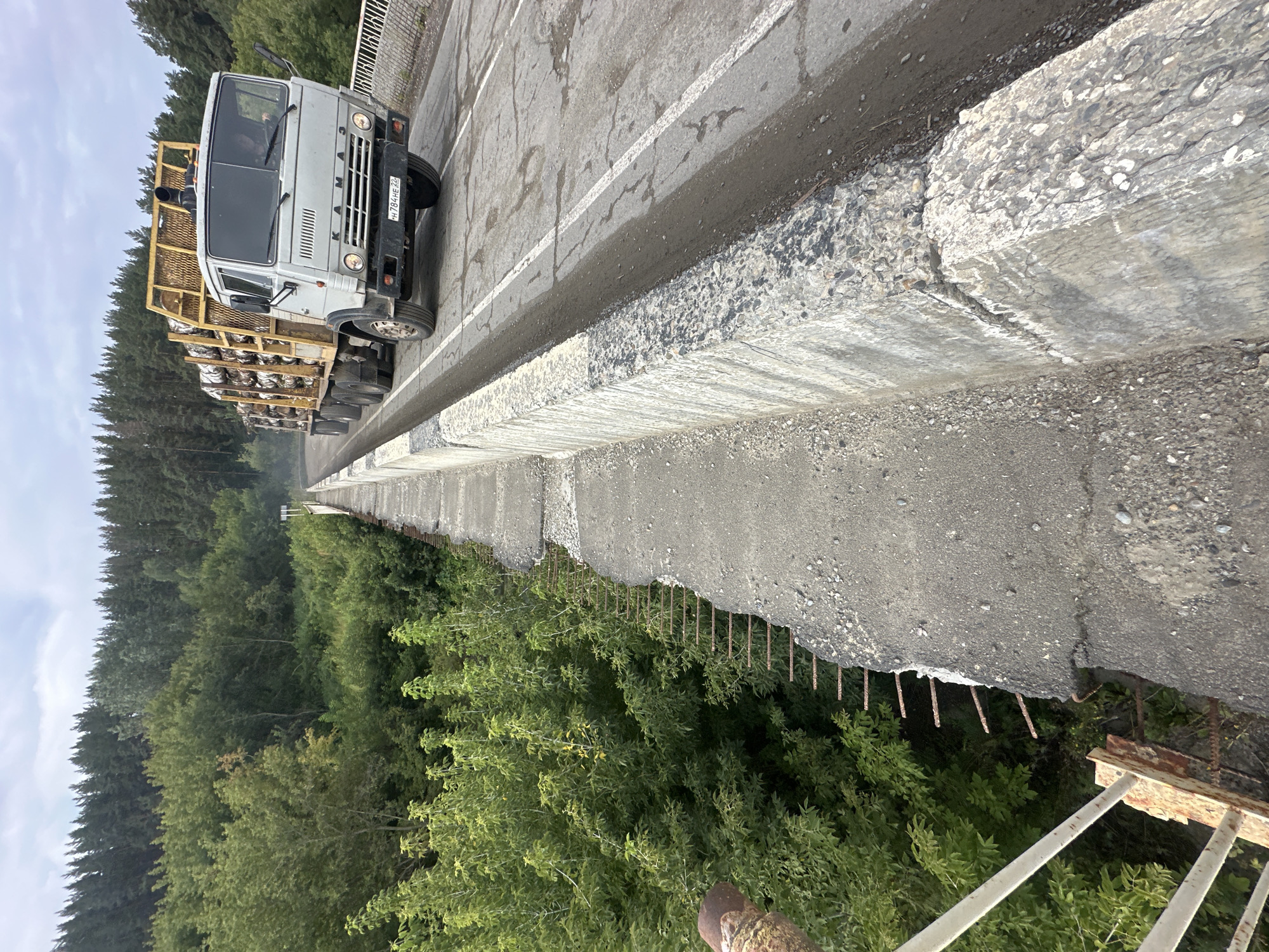 A light gray truck, identified as a KAMAZ by its badging, is driving across a weathered concrete bridge. The truck is loaded with what appear to be large wooden logs secured by a yellow metal frame. The bridge shows signs of disrepair, with crumbling concrete on the edges and exposed rebar. The road surface is cracked and dusty, with visible white lane markings. On either side of the bridge, dense green foliage, including trees and bushes, encroaches on the structure. The background features a thick forest of evergreen trees under a cloudy sky. The scene appears to be located in an outdoor, possibly rural, environment.