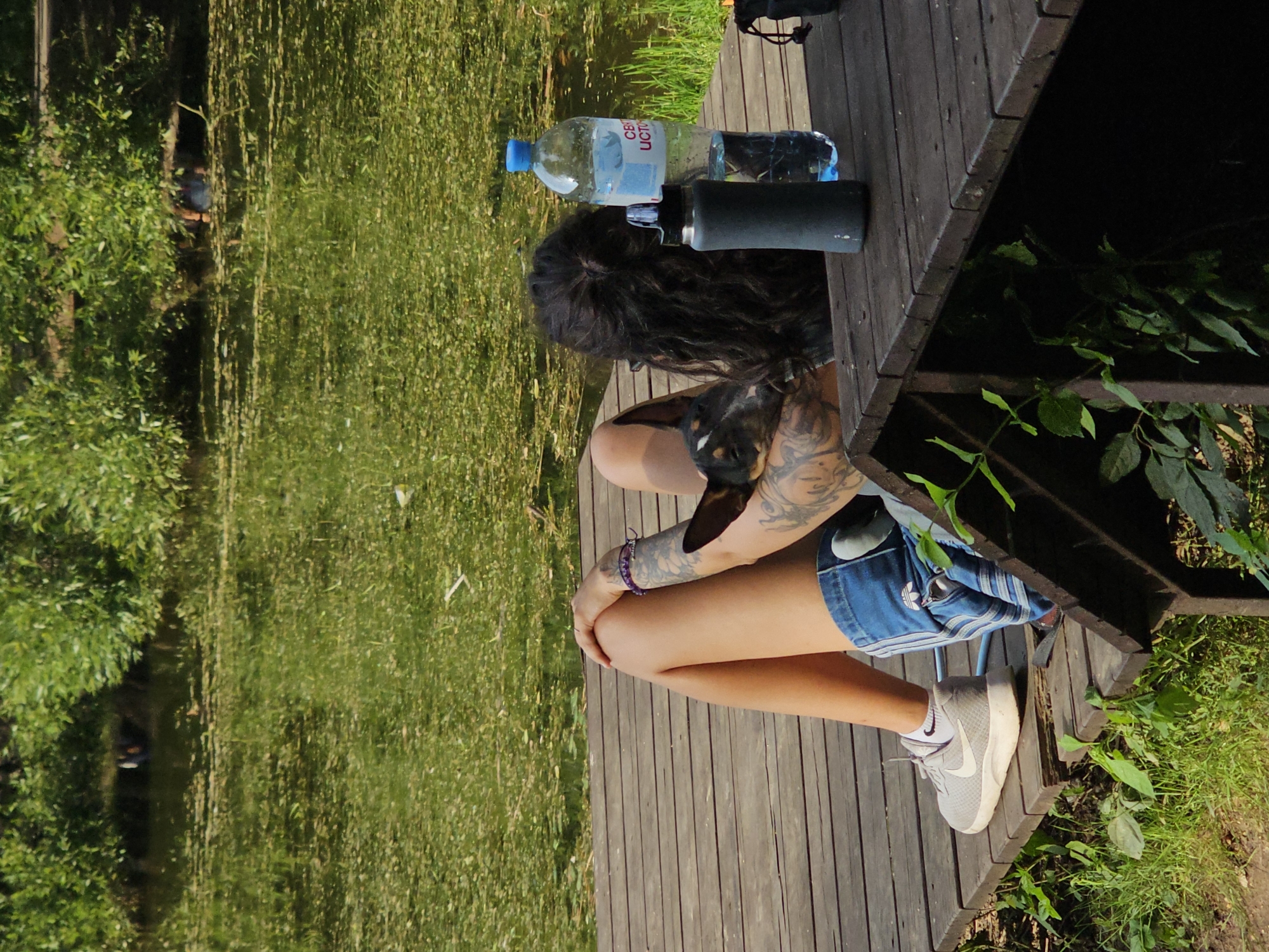 A person, with visible tattoos on their arm and leg, is sitting on a wooden deck next to a body of water. The person is wearing denim shorts and athletic shoes. A small dog rests on the person's arm. On the deck, to the right of the person, are two water bottles and a black thermal flask. The body of water is partially covered in green algae and surrounded by lush green foliage. The scene appears to be outdoors in a natural setting, possibly a park or a lakeside area within Moscow, Russia.