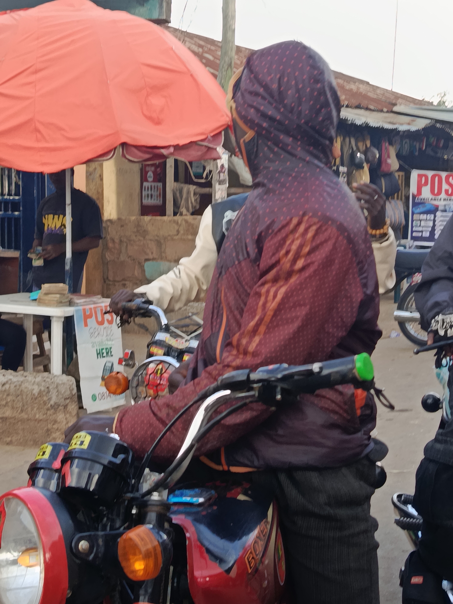 In Kabong, Nigeria, a street scene unfolds near a market stall. An individual wearing a hooded, patterned jacket sits on a red motorcycle, facing away from the camera. Their left hand rests on the motorcycle's handlebars, and their right arm is bent, partially obscuring their face. The motorcycle has a prominent headlight, orange turn signals, and the word "BOXER" visible on its side.
Behind the motorcycle rider, another person in a light-colored shirt is visible. To the left, a person in a dark shirt is standing by a white table laden with stacks of cash and what appears to be promotional flyers. One flyer on the table advertises "POS Service Available HERE" and shows images of a phone and a POS machine, along with a phone number and a green circular logo.
A large, bright orange umbrella provides shade over the area. In the background, market stalls with corrugated metal roofs are visible, displaying various goods. The ground is paved, and the overall impression is of a busy, informal commercial setting.