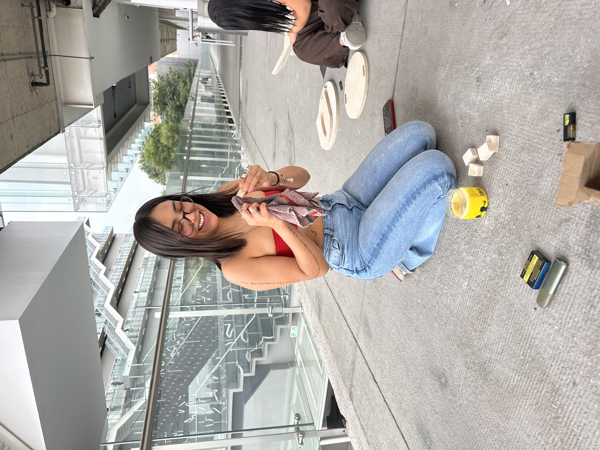 The image shows a woman in jeans and a red top sitting on a concrete surface, possibly a rooftop or terrace. She is holding a small wooden object and a cloth, seemingly involved in some kind of crafting or woodworking activity. Beside her are various crafting materials, including wooden blocks, a yellow container, and unfinished wooden shapes. Another person is seen in the background, also seated, and there are additional wooden shapes nearby. The location appears to be urban, with a modern building with glass panels and a stairway visible in the background. The building is likely in Mexico City, Mexico, as indicated in the prompt.