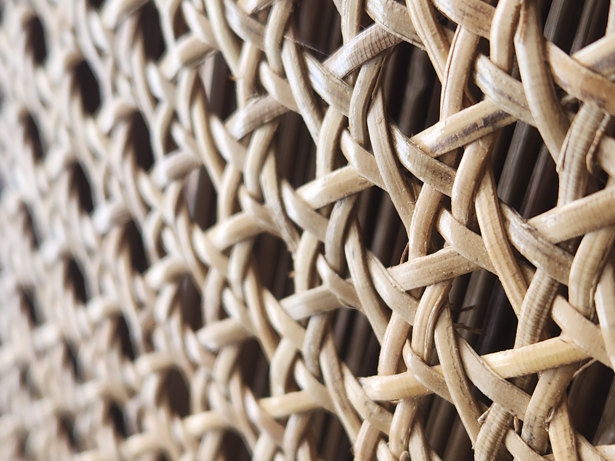 The image is a close-up, high-angle shot of a woven natural fiber material, possibly rattan or wicker. The fibers are light brown and arranged in an interlocking pattern creating a textured surface with open spaces. The depth of field is shallow, with the foreground in sharp focus and the background gradually blurring. The material appears to be part of a piece of furniture or decor. There are no people, animals, or visible indications of a specific location in the city of Maitland, United States of America within the image itself.