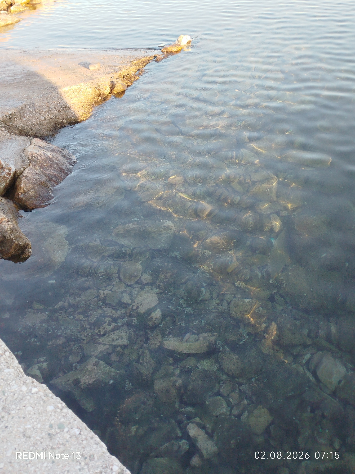 This image depicts a shallow body of clear water with a rocky seabed, adjacent to a concrete embankment. The water's surface shows gentle ripples and is illuminated by sunlight, which casts patterns on the submerged stones. A section of the concrete embankment is visible in the foreground, showing a rough texture and a few scattered rocks. In the distance, the water appears deeper and darker. The timestamp "02.08.2026 07:15" and "REDMI Note 13" are overlaid on the image, suggesting it was taken with a Redmi Note 13 phone on August 2, 2026, at 7:15 AM. Based on the metadata, the location is Sithonia Municipal Unit, Greece. There are no people or animals visible in the image. The scene is a natural coastal environment.