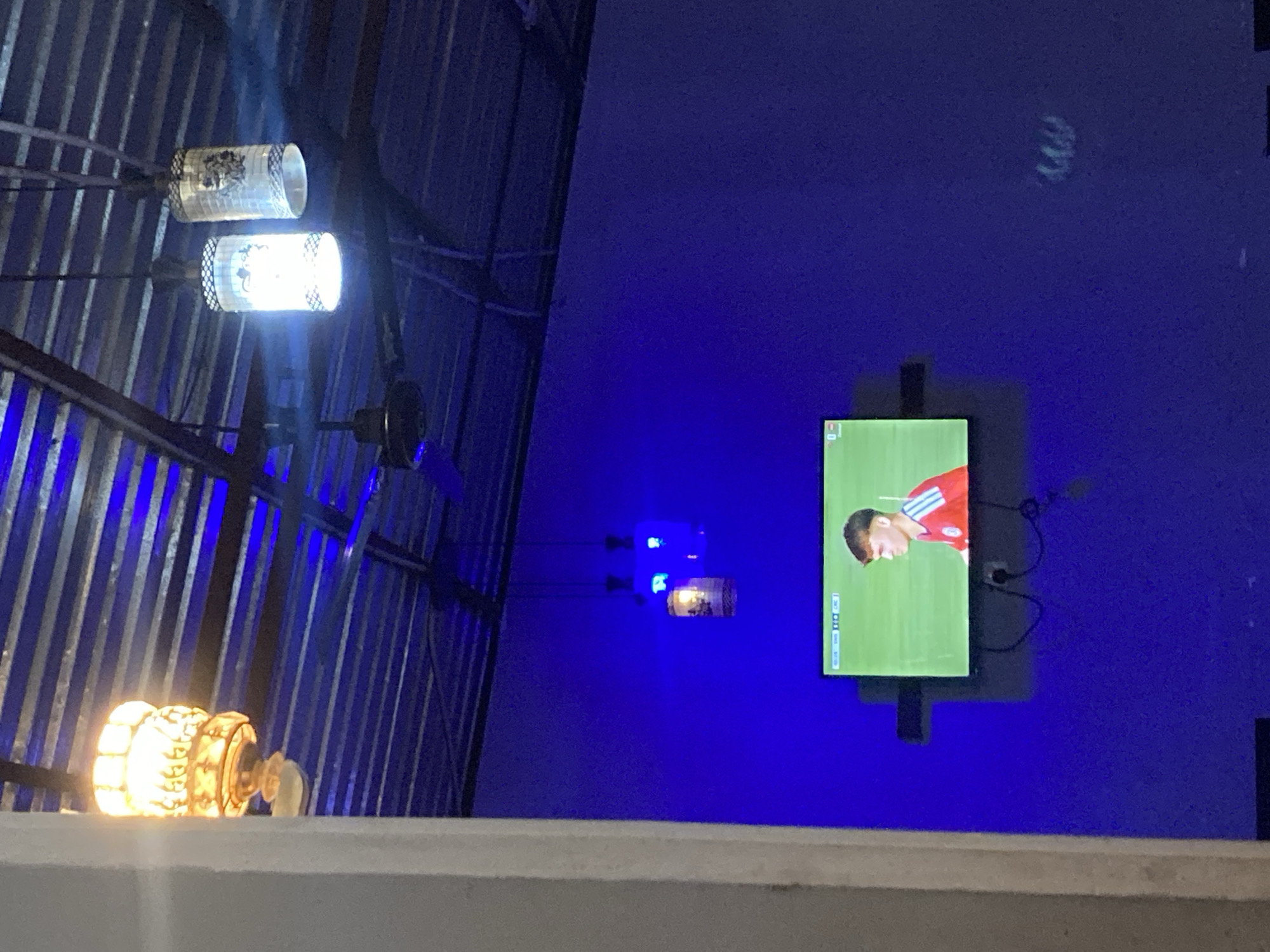 The image depicts an interior scene, likely a room or bar, with a television displaying a soccer match. The room is illuminated by blue lighting and features a corrugated metal ceiling with exposed beams and a ceiling fan. Hanging from the ceiling are three decorative lanterns, one with an ornate, golden design and two cylindrical white lanterns with patterned detailing. A white pillar is visible in the foreground on the left side of the frame. The television, mounted on the wall, shows a young man in a red soccer jersey with white stripes on the sleeves. The overall ambiance is dimly lit, with the blue lights creating a vibrant and somewhat atmospheric setting. This scene is located in Gwarinpa, Nigeria.