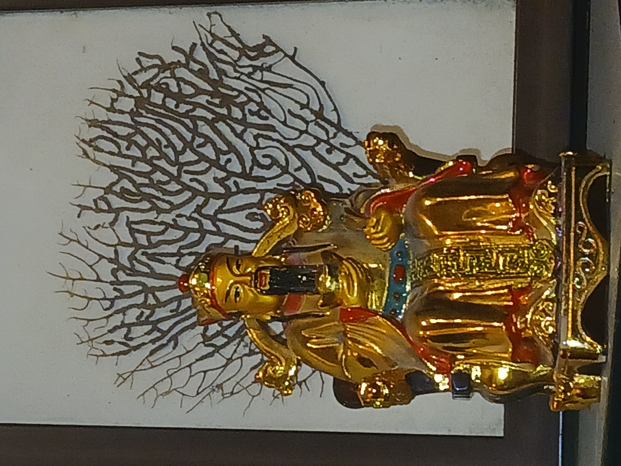The image shows a golden statue of a deity placed against a framed picture. The picture has a white background with a brown, tree-like pattern on it. The deity is holding a dark object in his hand and is seated on an ornate throne. The photo appears to have been taken in a dimly lit indoor environment. Given the likely cultural context of such iconography, it may be assumed the location is Bengaluru, India.