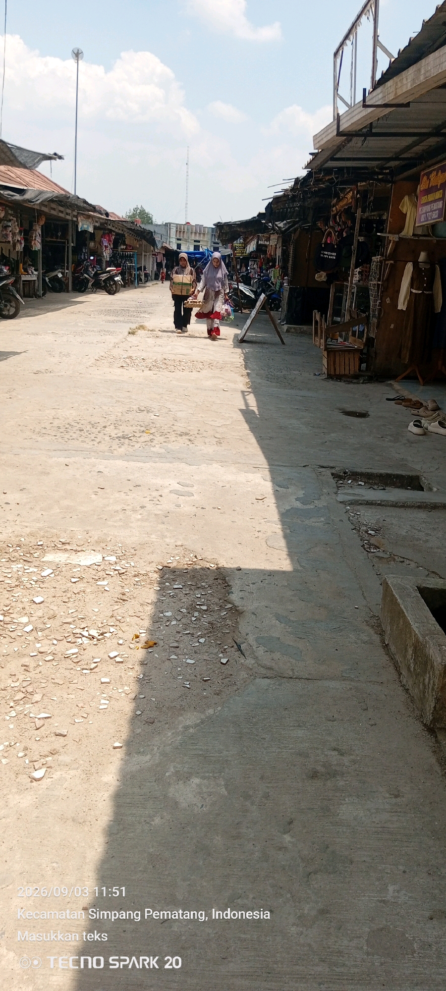 Two women are walking down a dirt and concrete road lined with market stalls. The woman on the left is wearing a yellow headscarf and dark pants, carrying a box. The woman on the right wears a light purple headscarf and a patterned dress, carrying a woven basket. The market stalls on both sides display various goods, with some motorcycles parked nearby. The sky is bright and partly cloudy. A timestamp and location are visible at the bottom of the image: "2026/09/03 11:51 Kecamatan Simpang Pematang, Indonesia".