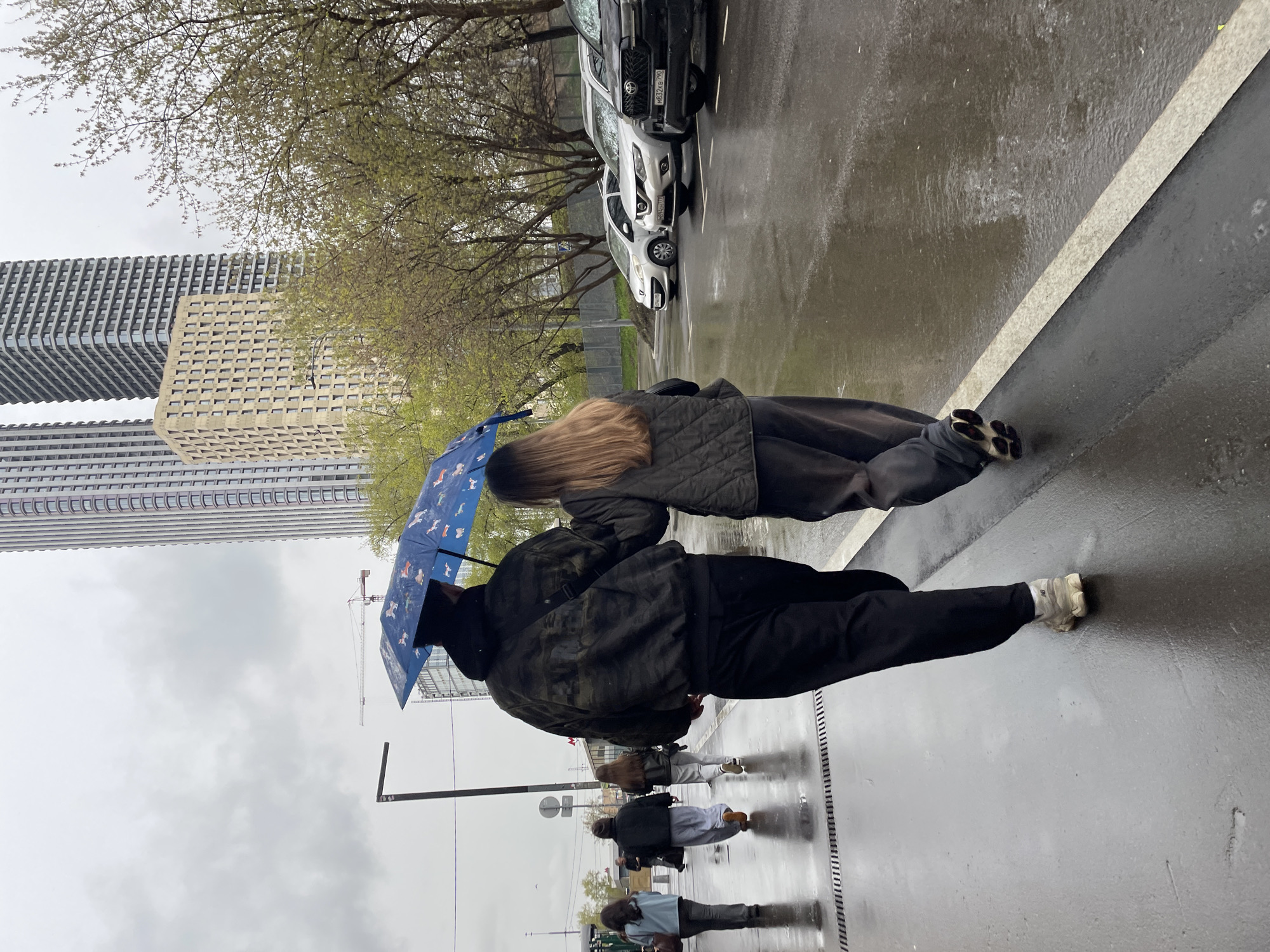 On a wet city street in Khimki, Russia, a man and a woman share a blue umbrella while walking away from the camera. They are dressed in dark clothing and appear to be a couple. Other pedestrians walk in the opposite direction, their reflections visible on the wet pavement. In the background, there are modern buildings, parked cars, and bare trees. The sky is overcast and gray.