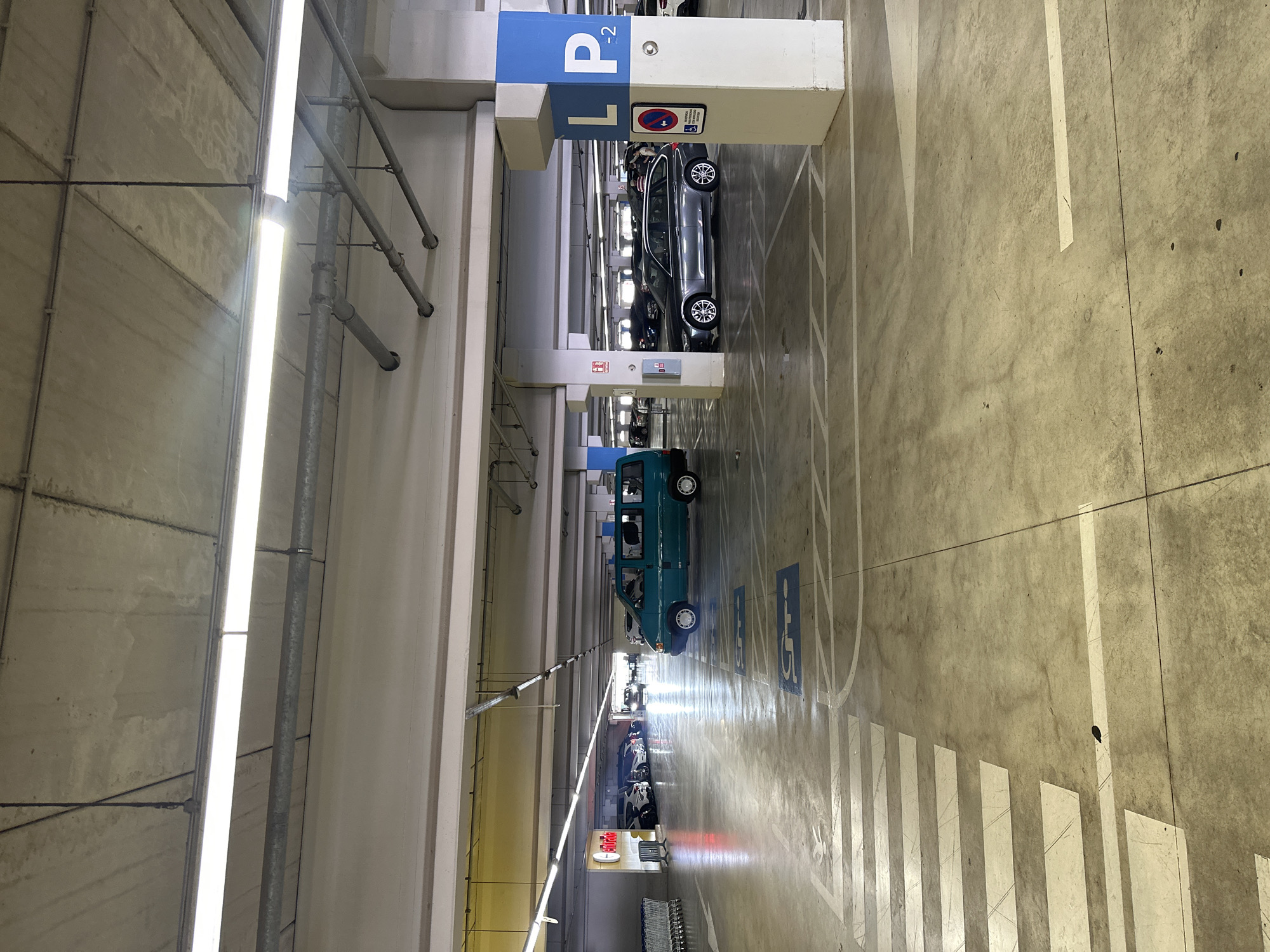 This is a photograph taken in a multi-story parking garage, likely in Alfafar, Spain, as indicated by the location context. The perspective is from above, looking down into the garage. The scene is illuminated by long fluorescent lights running parallel to the rows of parking spaces.
The primary subjects are cars parked in their designated spots. A grey sedan is visible in the upper portion of the image, parked under a blue and white sign that reads "P L2". A teal-colored van is parked further down the row, and several other vehicles are visible receding into the distance. The floor of the garage is concrete, marked with white lines indicating parking bays and directional arrows. There are also blue symbols on the floor indicating disabled parking spaces.
The setting is an indoor parking facility, characterized by concrete pillars, overhead lighting, and a general utilitarian aesthetic. There are no visible people in the frame, suggesting it might be a quiet time. The time of day cannot be determined from the image, and the weather is irrelevant as it is an indoor setting.
Notable details include the signs indicating parking levels and accessibility, as well as the reflective surface of the floor, likely from a polished concrete finish or a light coating. There are also visible pipes and conduits running along the ceiling and walls, typical of infrastructure in such a building. A red and white sign with text, possibly a brand or advertisement, is visible further down the row. The overall impression is one of order and functionality.